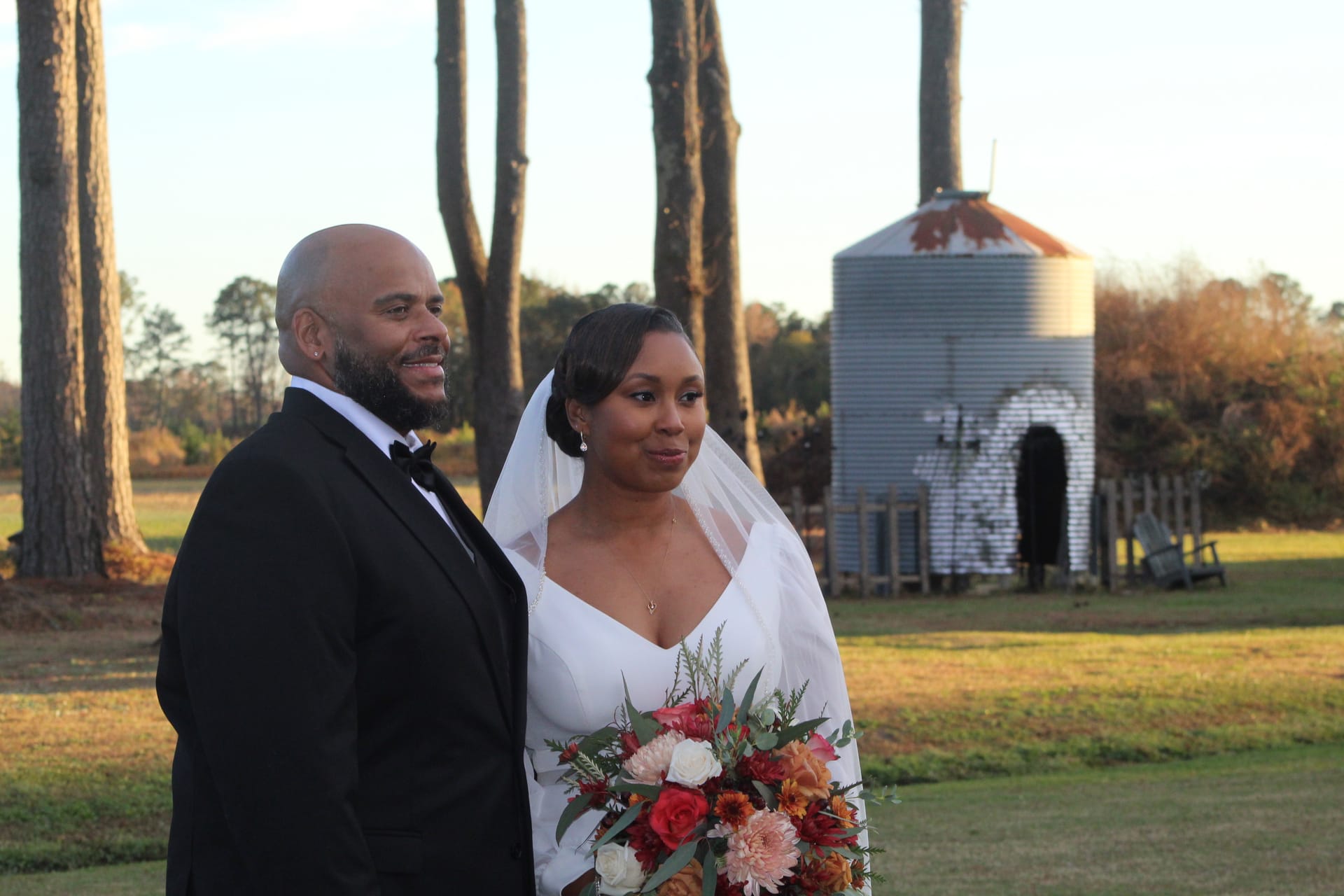 Bride in white gown holding a colorful autumn bouquet stands beside her tuxedo-clad groom on the open farmland grounds of Scarborough House with a rustic grain silo in the background at golden hour.