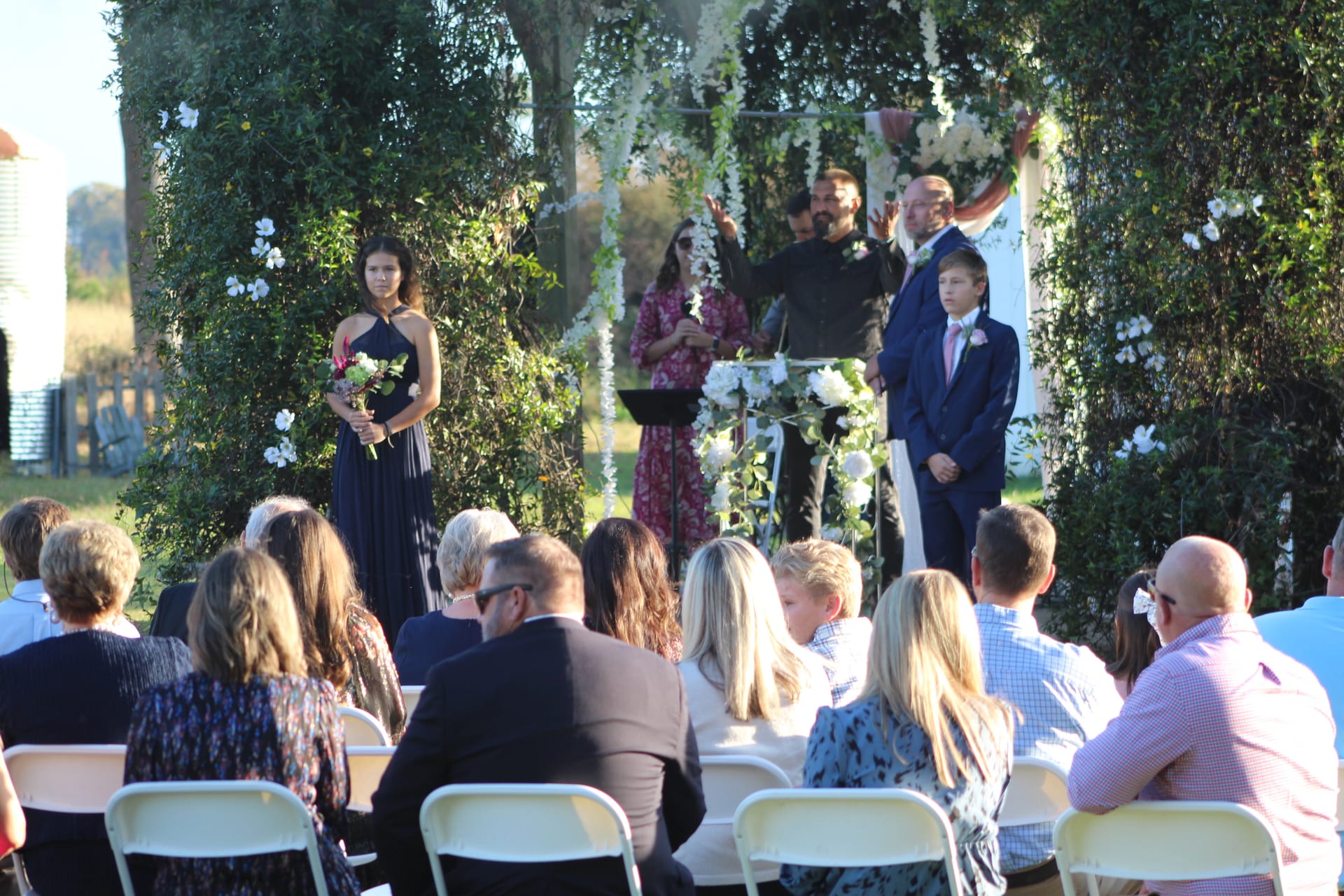 Outdoor wedding ceremony on the estate lawn with guests seated as the officiant speaks beneath a white floral arch surrounded by lush greenery and farmland fields.