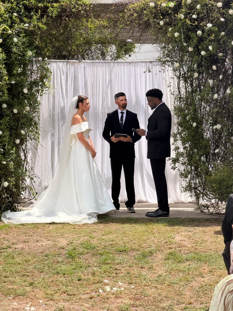 Bride in an off-the-shoulder ball gown and veil exchanges vows with her groom before an officiant beneath a lush floral arch with white draped backdrop on the ceremony lawn at Scarborough House.