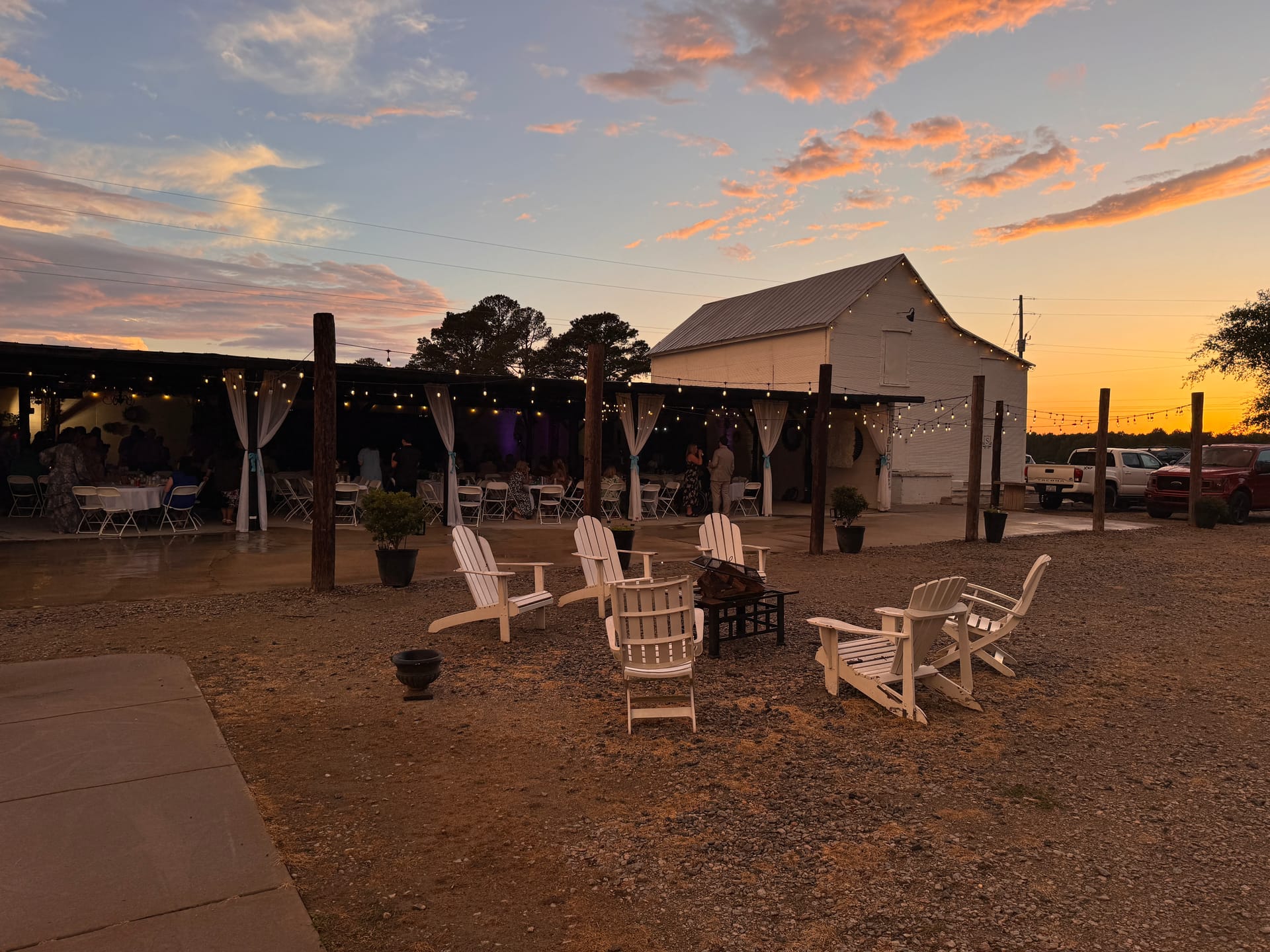 White Adirondack chairs arranged around a fire pit at dusk with string lights and a barn reception venue glowing in the background under a dramatic orange and pink sunset sky.