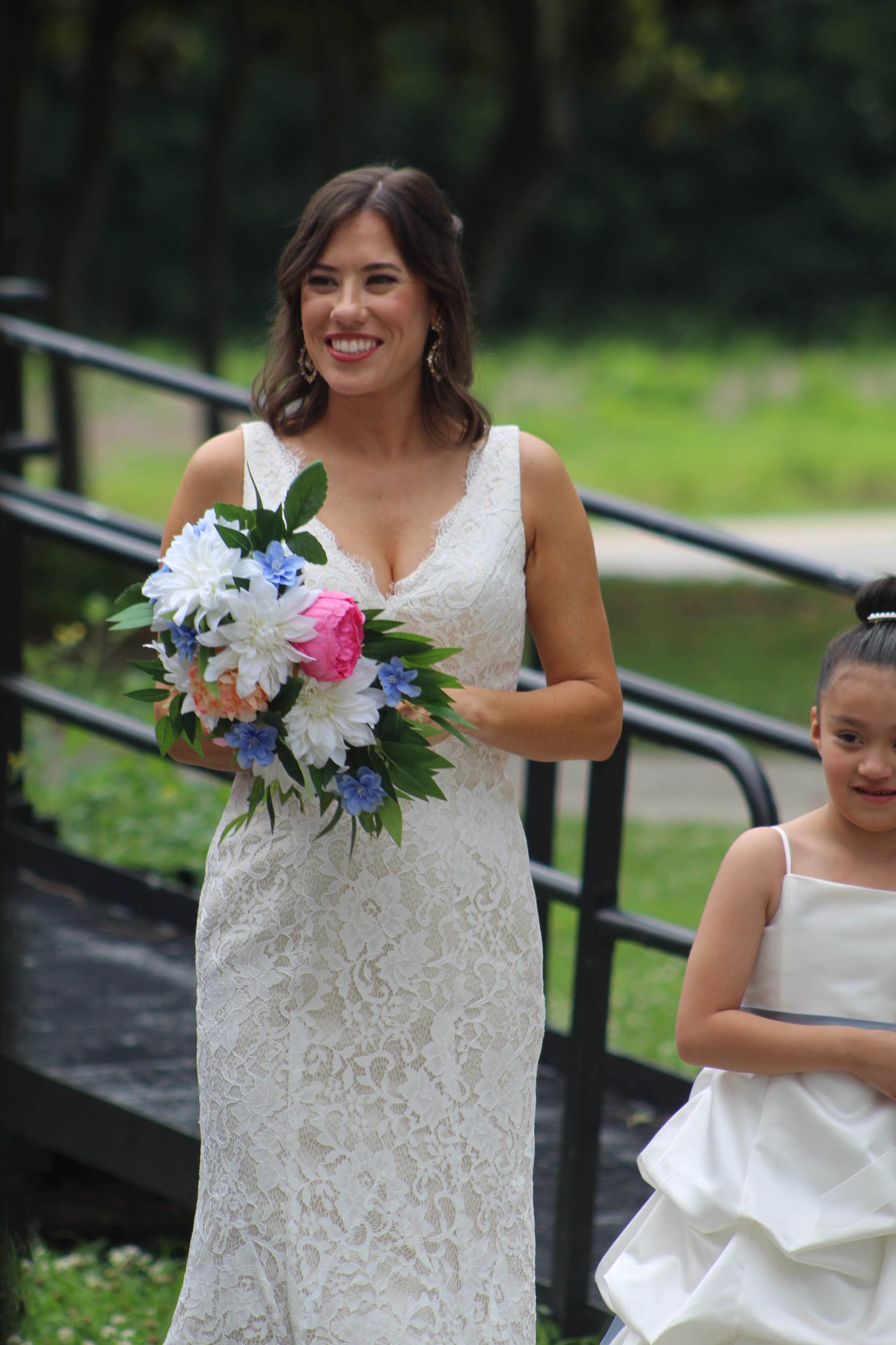 Smiling bride in a lace wedding gown holding a bouquet of pink peonies and blue flowers descends outdoor steps alongside a young flower girl at Scarborough House.