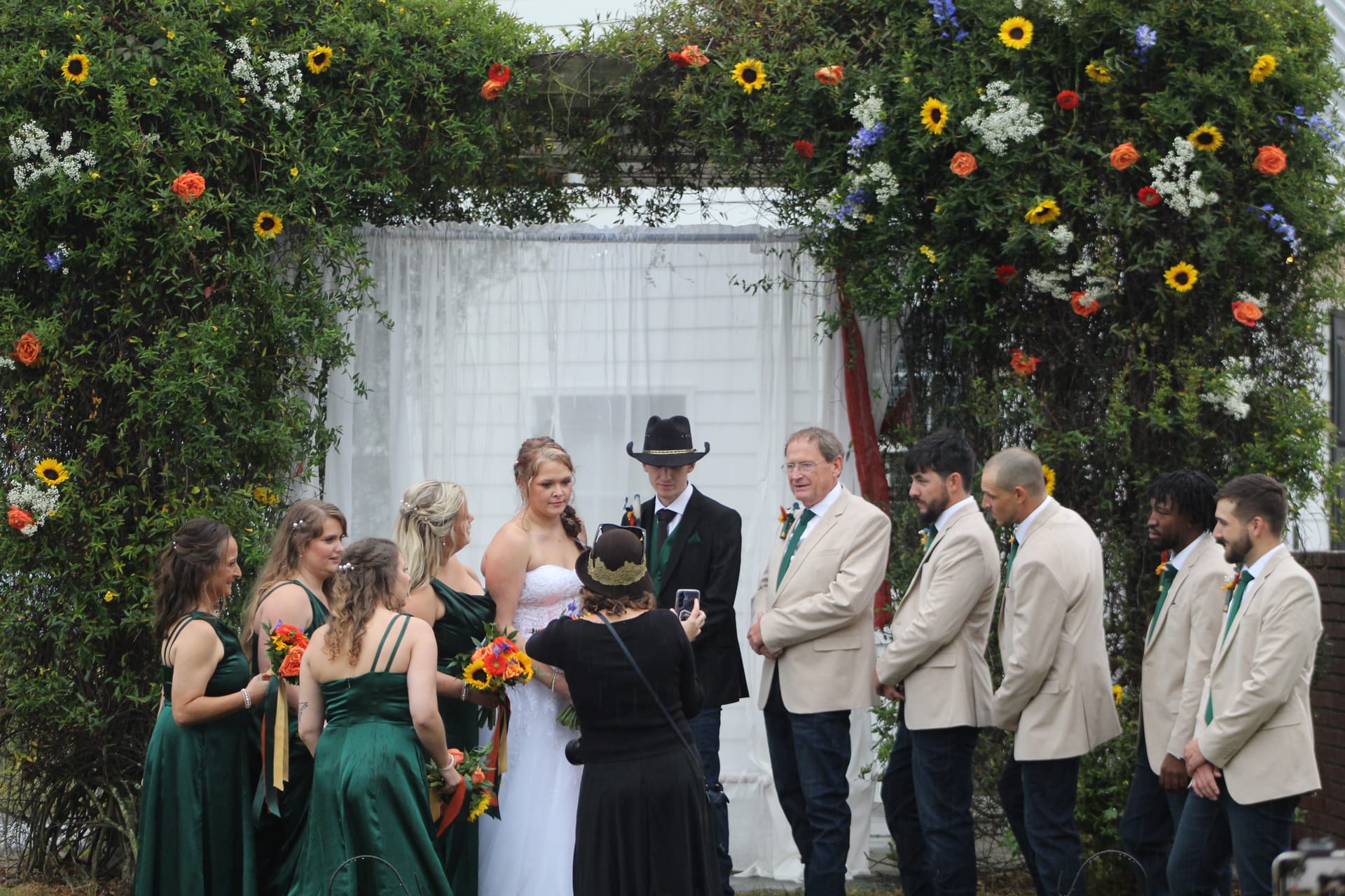 Wedding party gathered under a lush green floral arch adorned with sunflowers and orange roses during an outdoor ceremony at Scarborough House historic estate.