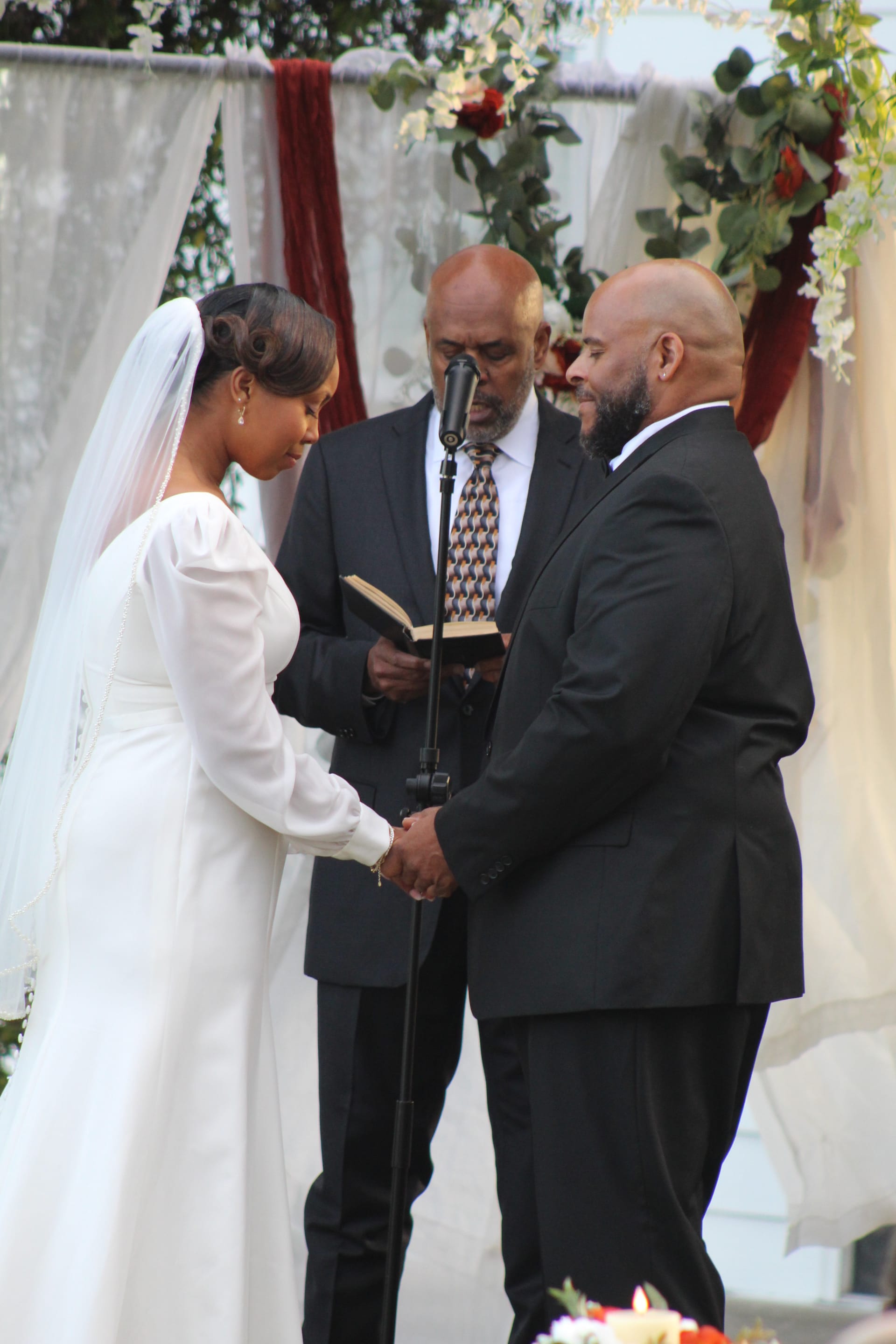 Bride in a long-sleeve white gown and veil holds hands with her groom in a black suit as an officiant reads from a book beneath a floral arch decorated with red and white blooms and greenery during an outdoor wedding ceremony.