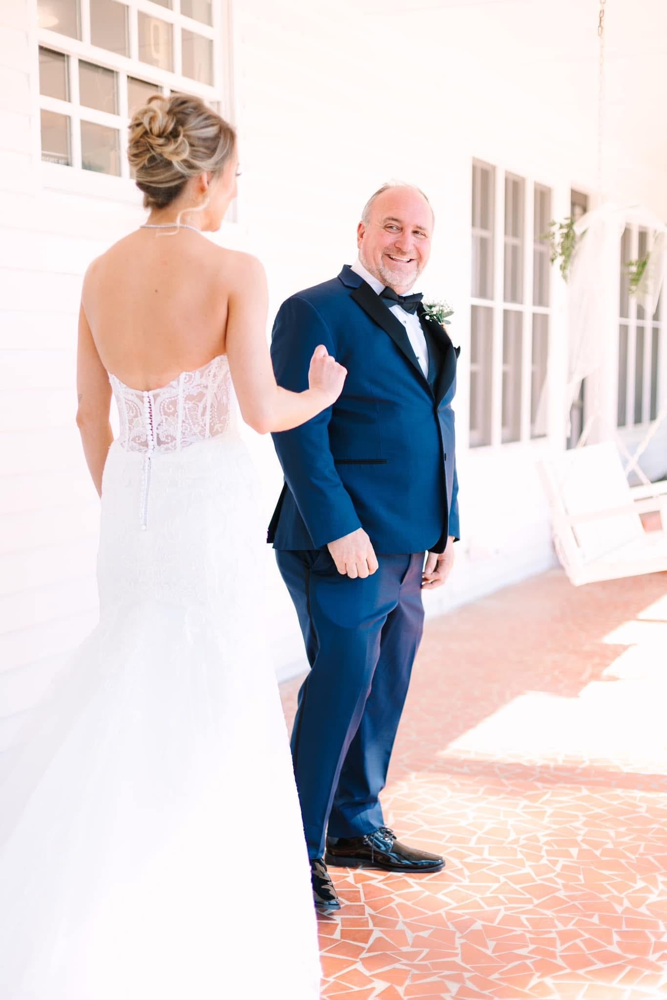 Groom in a navy tuxedo beams with joy as he turns to see his bride in a strapless lace gown during their first look on the sunlit porch of Scarborough House.