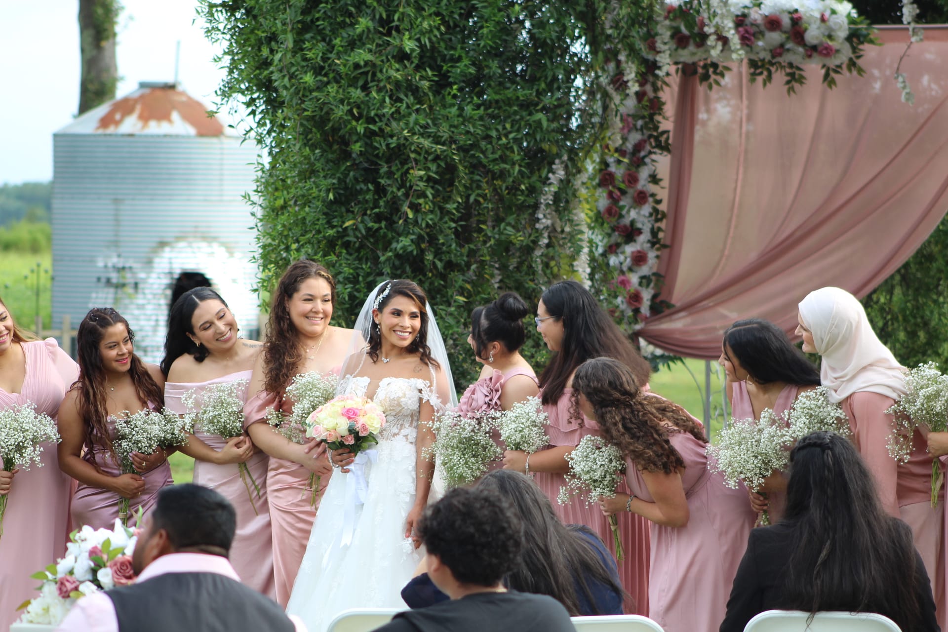 A radiant bride in a white lace gown walks down the aisle surrounded by smiling bridesmaids in blush pink dresses holding baby's breath bouquets beneath a floral and draped arch at Scarborough House.