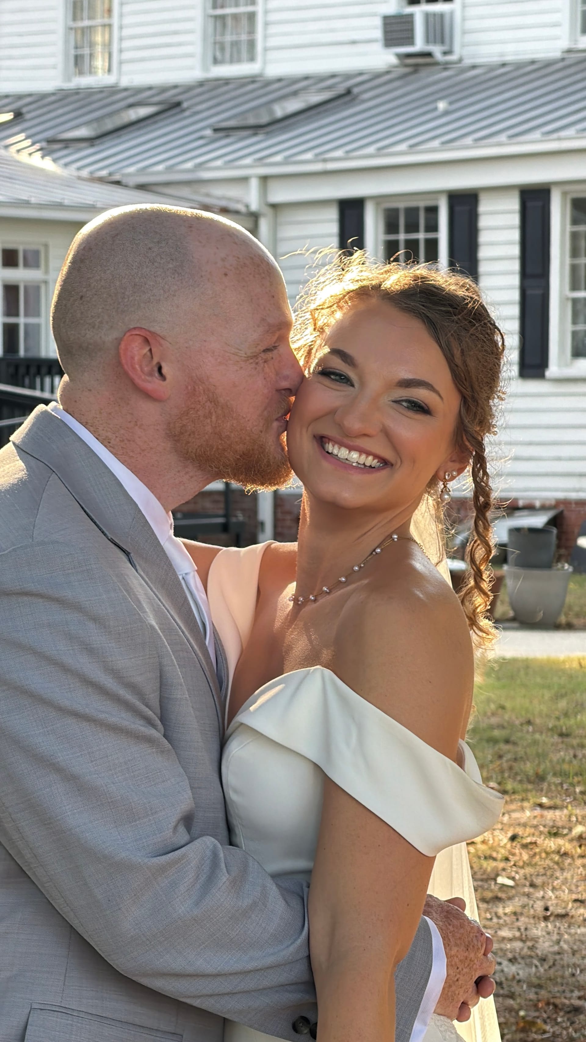 Groom in a light grey suit kisses the cheek of his smiling bride in an off-the-shoulder white gown during golden-hour portraits on the grounds of a historic 1821 estate wedding venue.