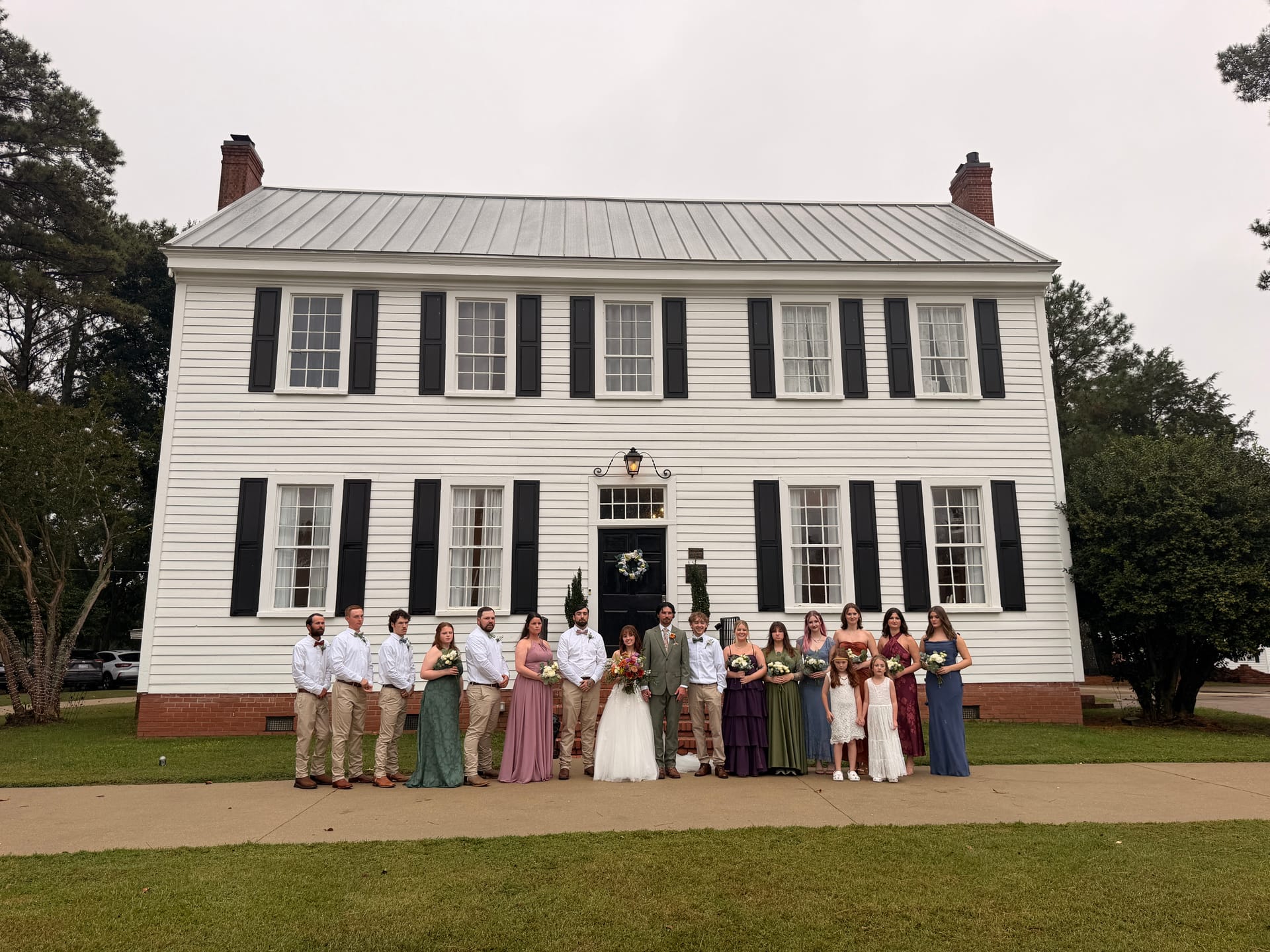 Full wedding party posing in front of the white two-story 1821 Scarborough House manor with black shutters on an overcast day.