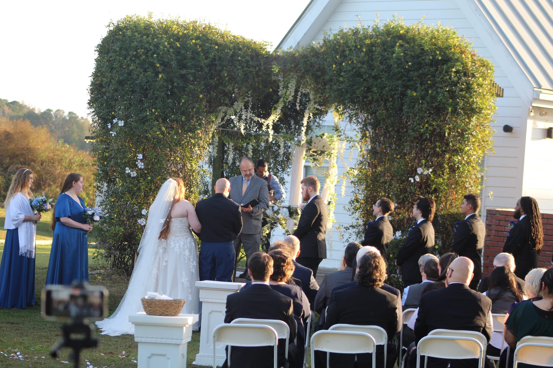 Bride and groom exchange vows beneath a lush ivy arch adorned with white florals on the ceremony lawn at Scarborough House historic estate, surrounded by wedding party and seated guests in golden afternoon light.