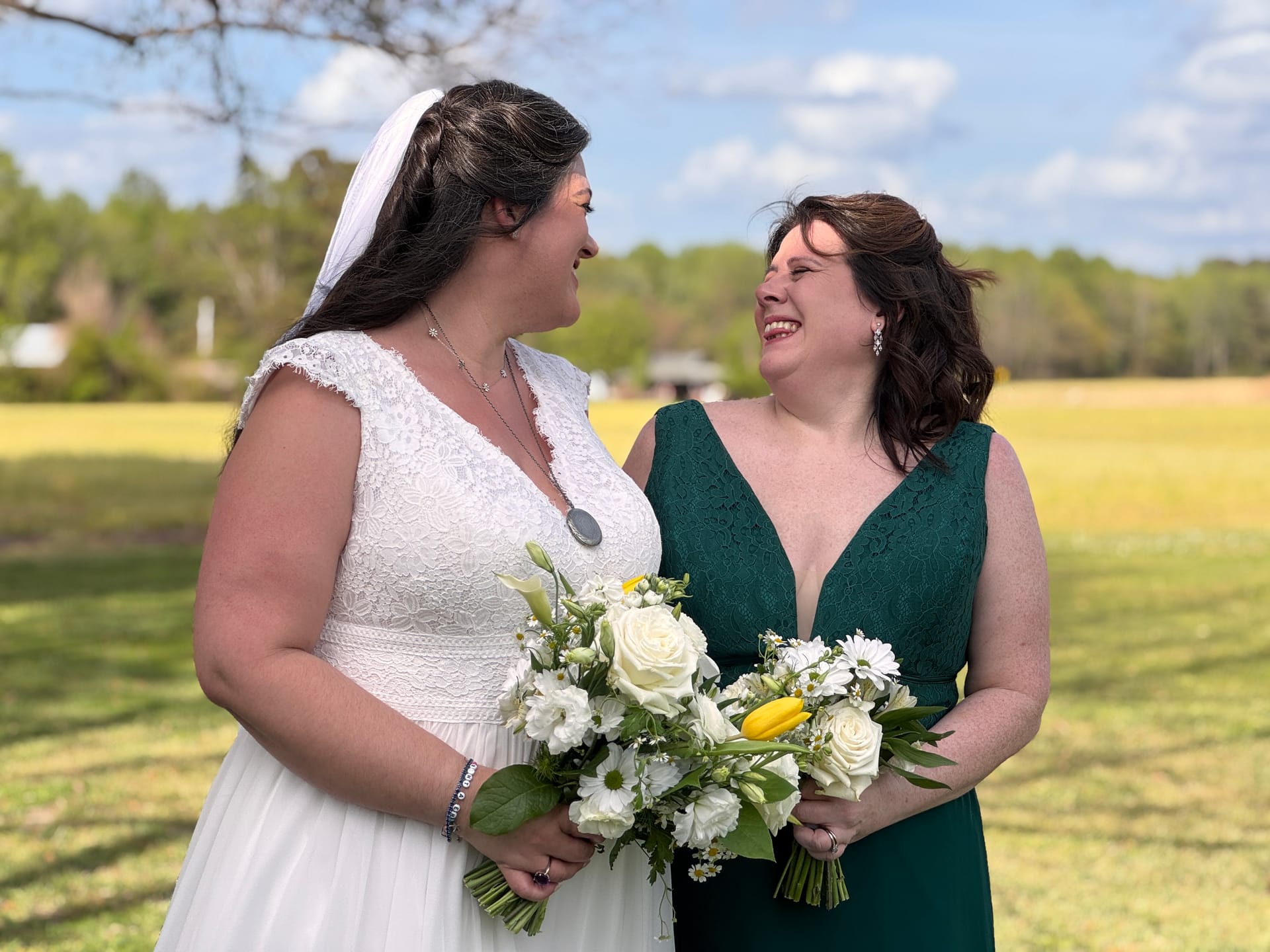 Bride in white lace gown and veil shares a joyful glance with her bridesmaid in an emerald green dress, both holding white and yellow bouquets against open farmland fields and blue sky at Scarborough House.