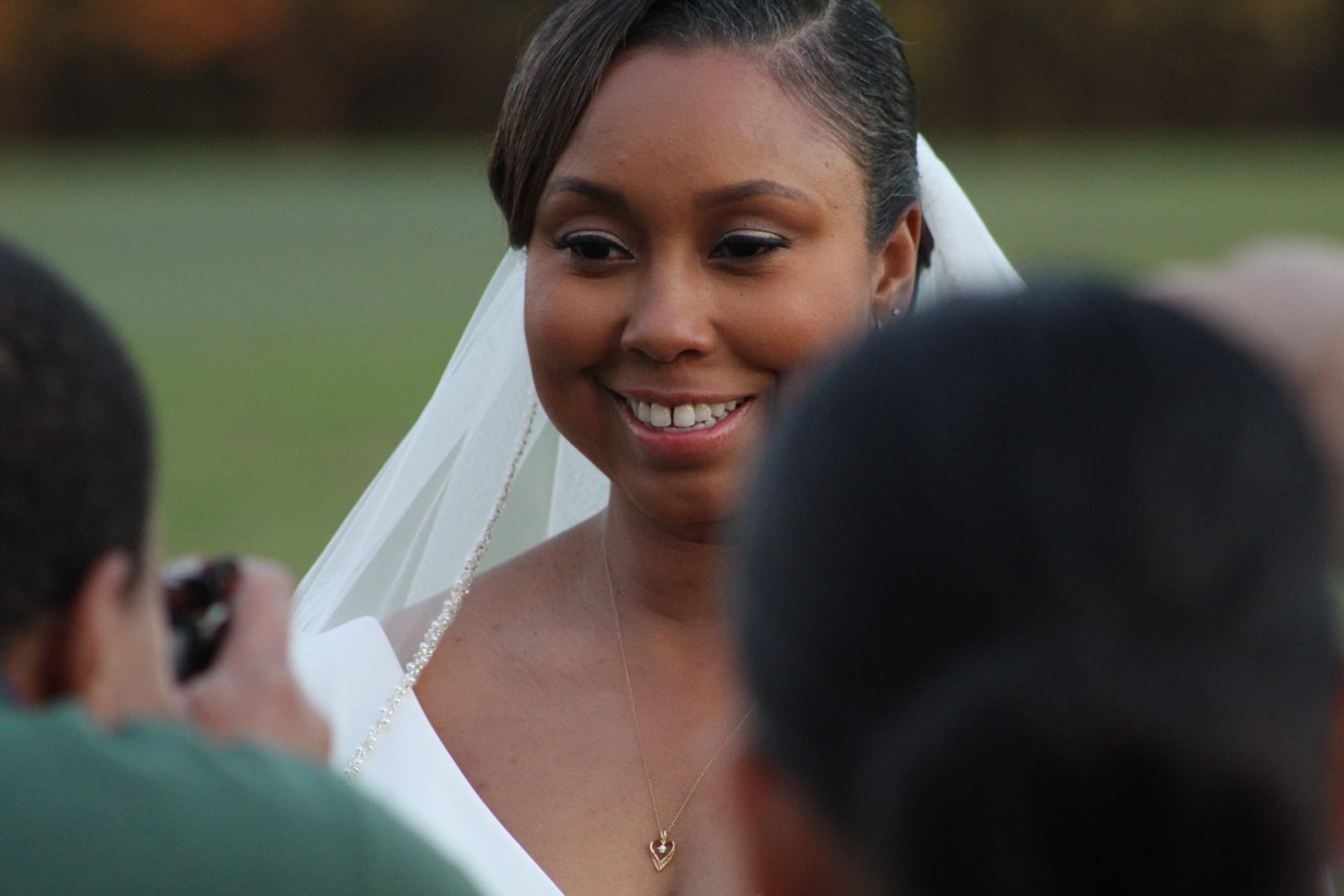 A smiling bride wearing a white veil and gold necklace stands outdoors on the ceremony lawn at Scarborough House estate venue.