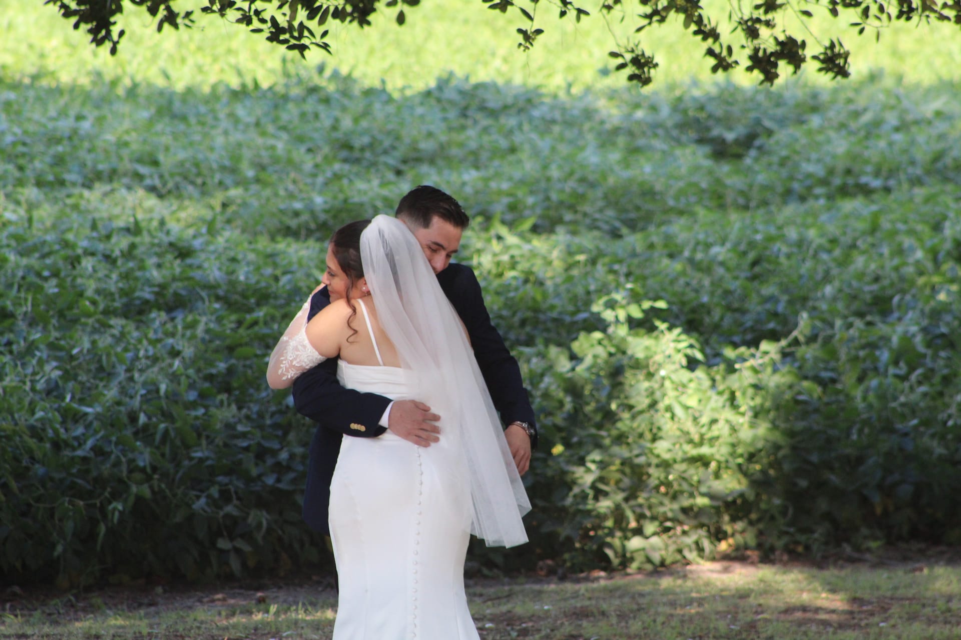 Bride in a fitted white gown and veil embraces her groom in a navy suit during an intimate first-look moment surrounded by lush green gardens at Scarborough House.