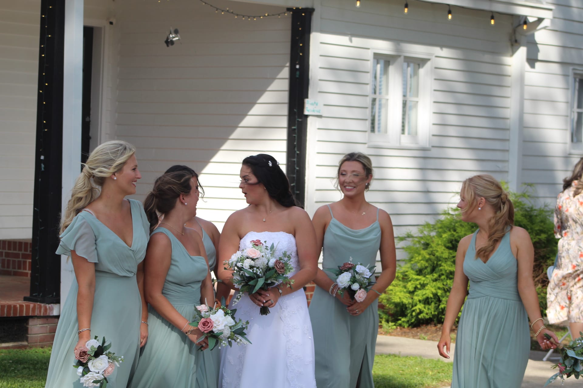 Bride in a strapless white gown holding a blush and white bouquet laughs with four bridesmaids in sage green dresses outside the historic white clapboard Scarborough House.