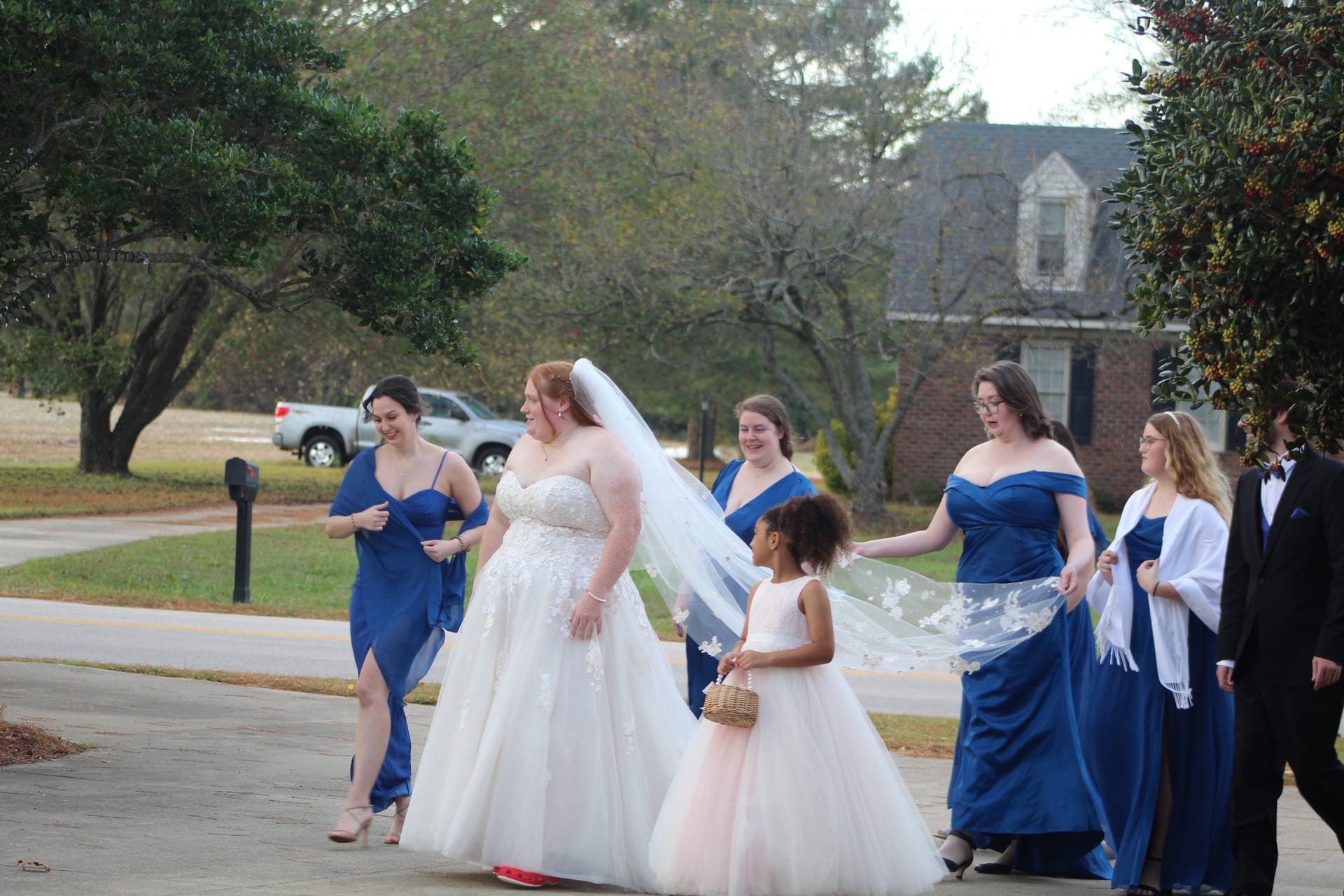 Bride in a white lace ballgown walks with her bridesmaids in royal blue dresses and a flower girl carrying a basket on a tree-lined path at Scarborough House.