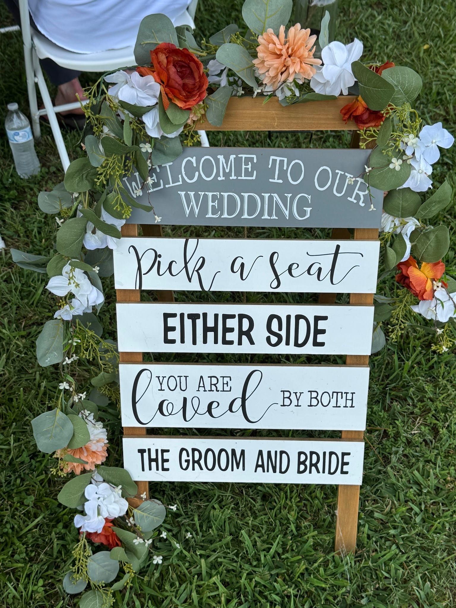 A wooden wedding welcome sign adorned with orange and white floral garland reads Pick a Seat Either Side You Are Loved by Both the Groom and Bride on a green lawn at Scarborough House.