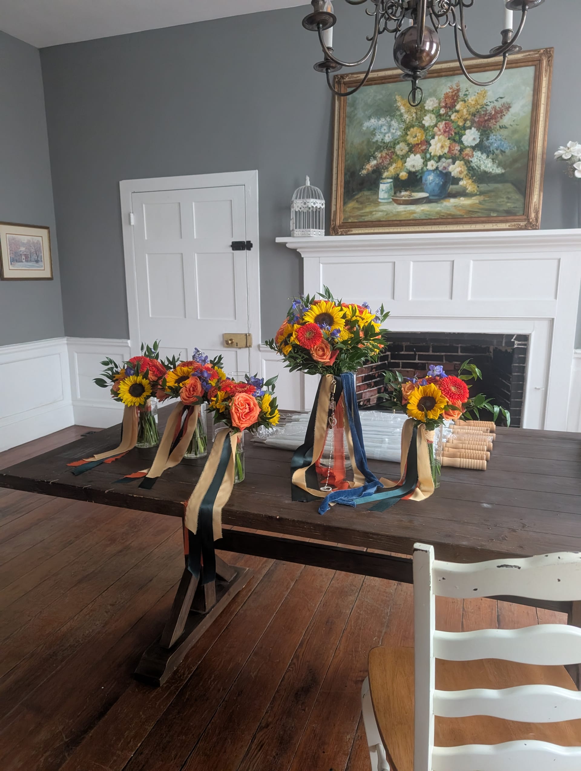 Colorful sunflower and rose bridal bouquets with navy and gold ribbons arranged on a dark wood farmhouse dining table inside the historic Scarborough House grand hall, with a floral oil painting above the white fireplace mantle.