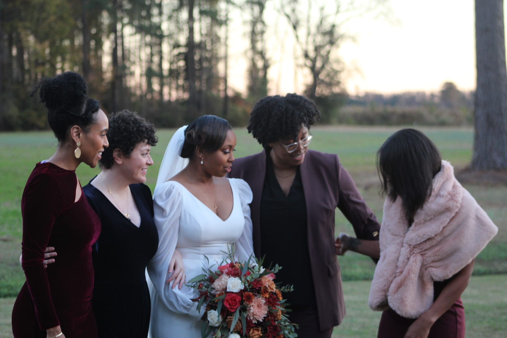 Bride in white gown with veil holding a fall floral bouquet shares a joyful candid moment with four bridesmaids on the open fields of Scarborough House at sunset.