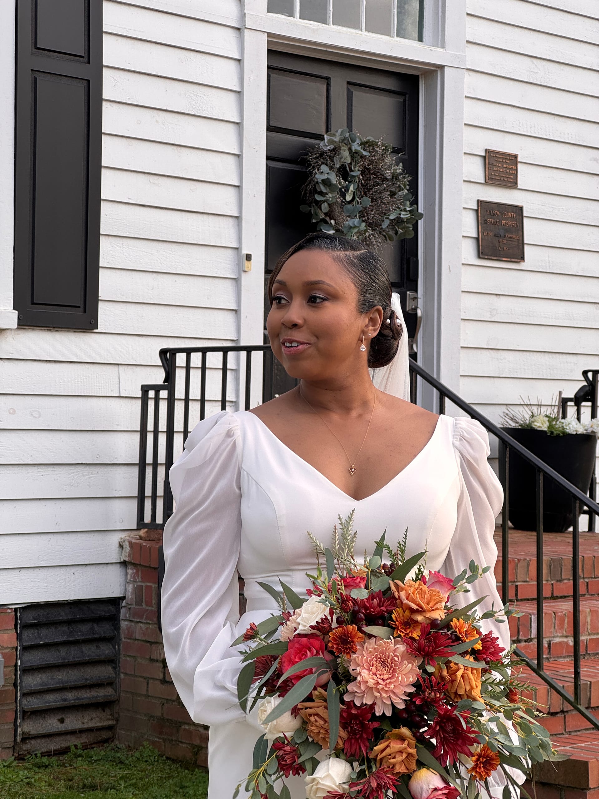 Bride in a long-sleeve white gown holds a cascading bouquet of burgundy, orange, and blush flowers on the front steps of the 1821 Scarborough House historic manor.