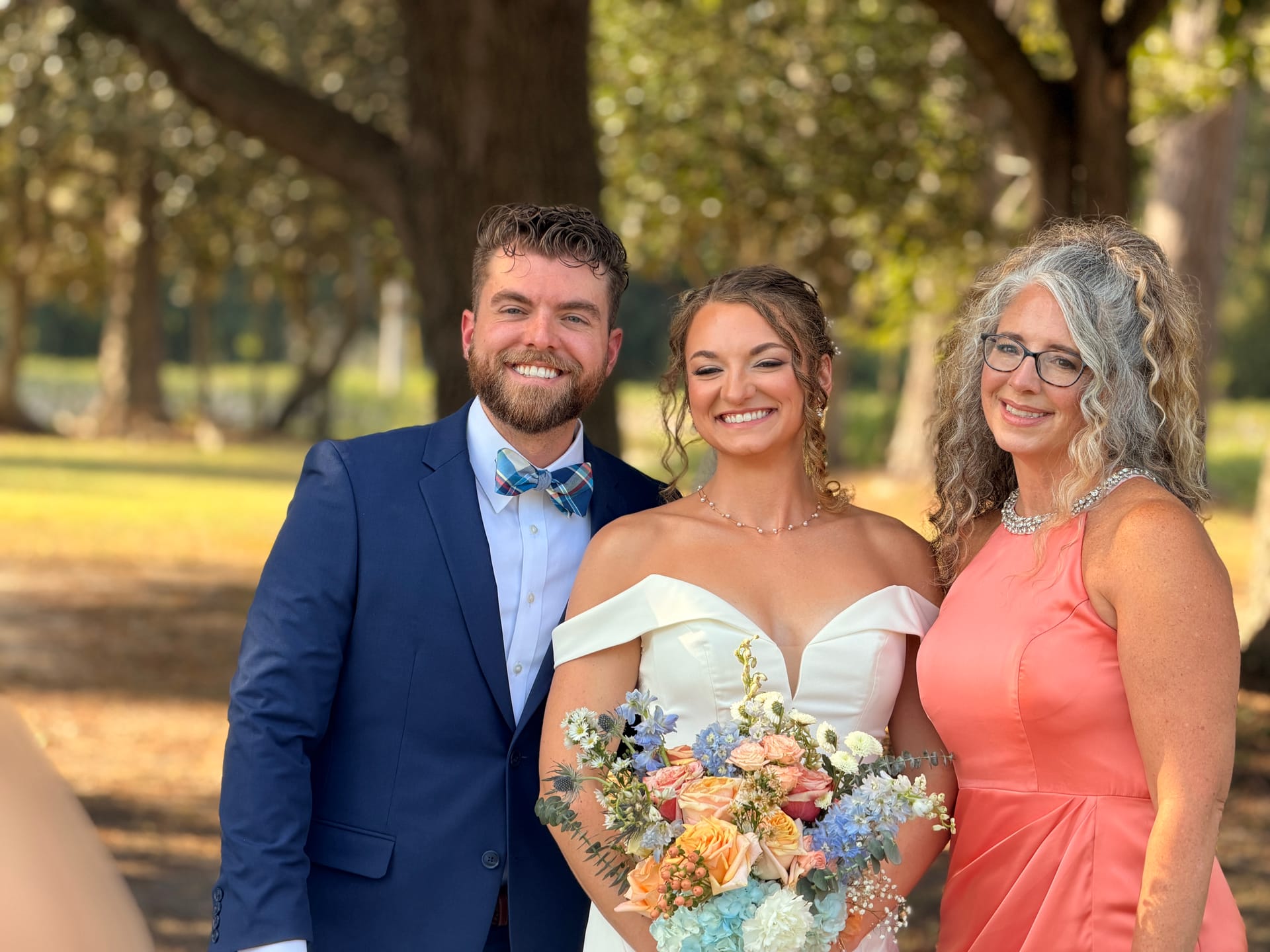 Bride in an off-the-shoulder white gown holding a pastel bouquet poses with the groom in a navy suit and plaid bow tie and an older woman in coral dress beneath the oak trees at Scarborough House.