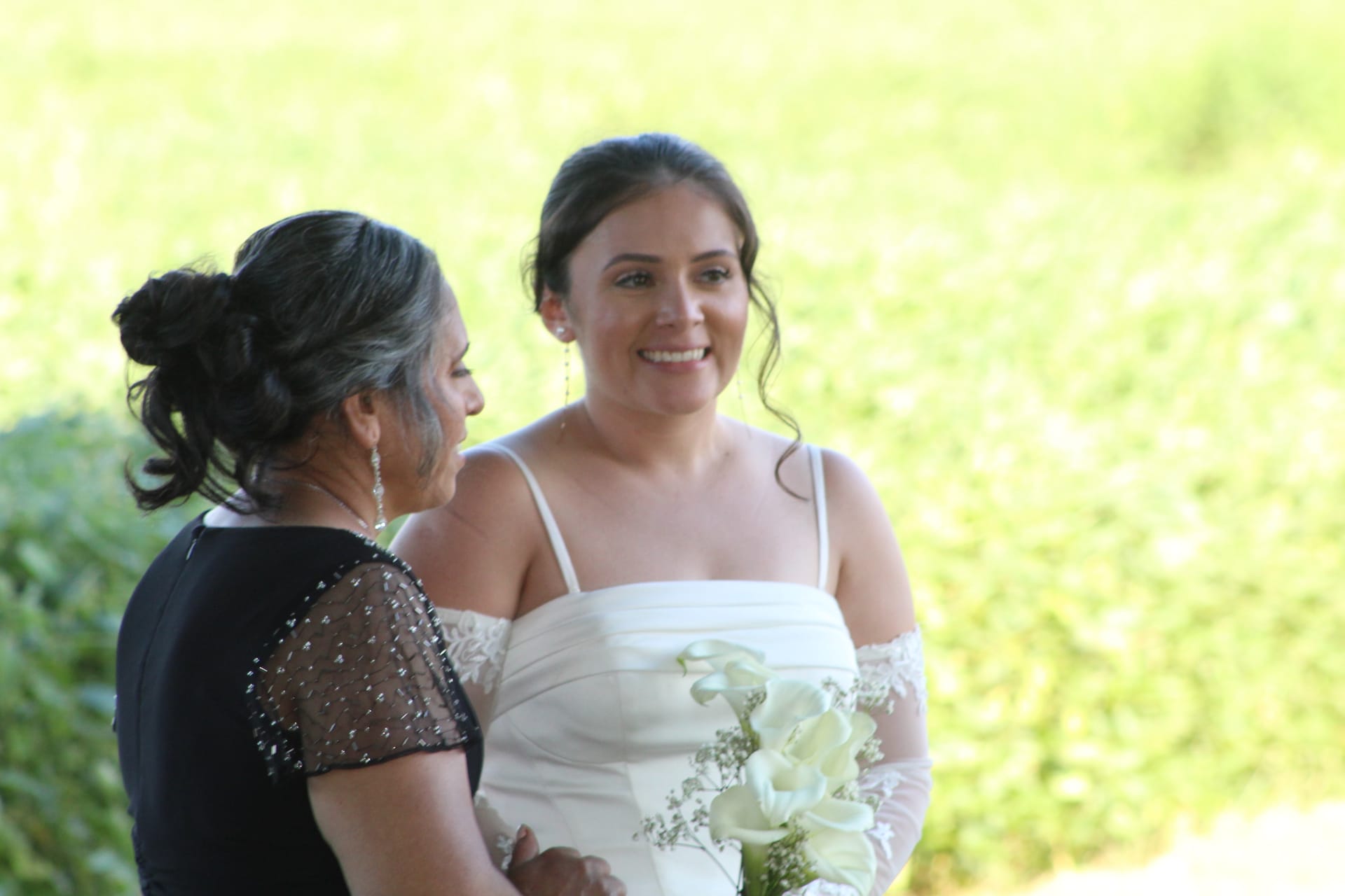 Smiling bride in a white off-shoulder gown holding white calla lilies shares a tender moment with an older woman in a black beaded dress against a lush green garden backdrop at Scarborough House.