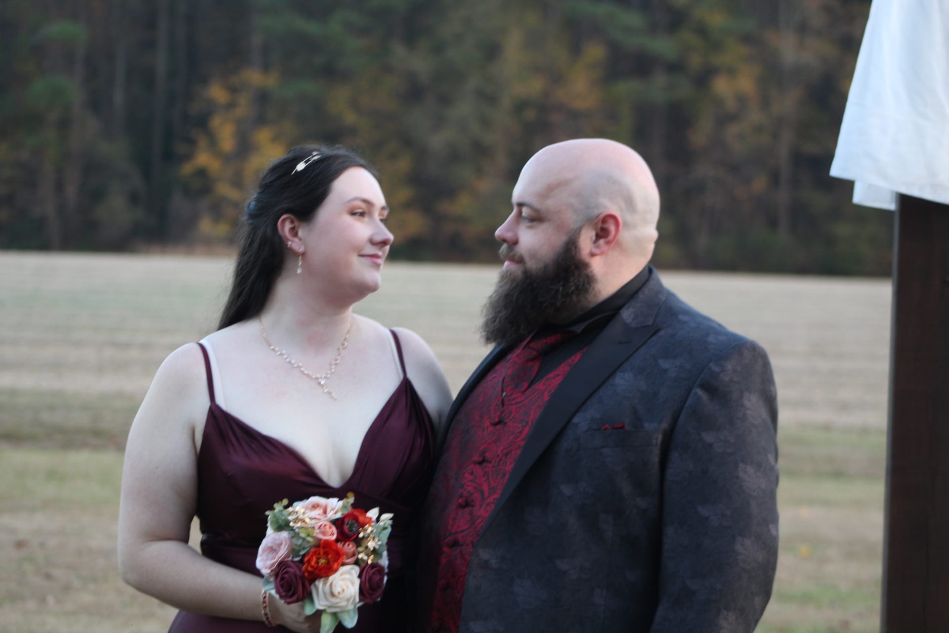 Bride in a burgundy gown holding a red and blush bouquet exchanges a loving glance with her bearded groom in a dark floral suit and crimson tie during an outdoor ceremony on the estate lawn.
