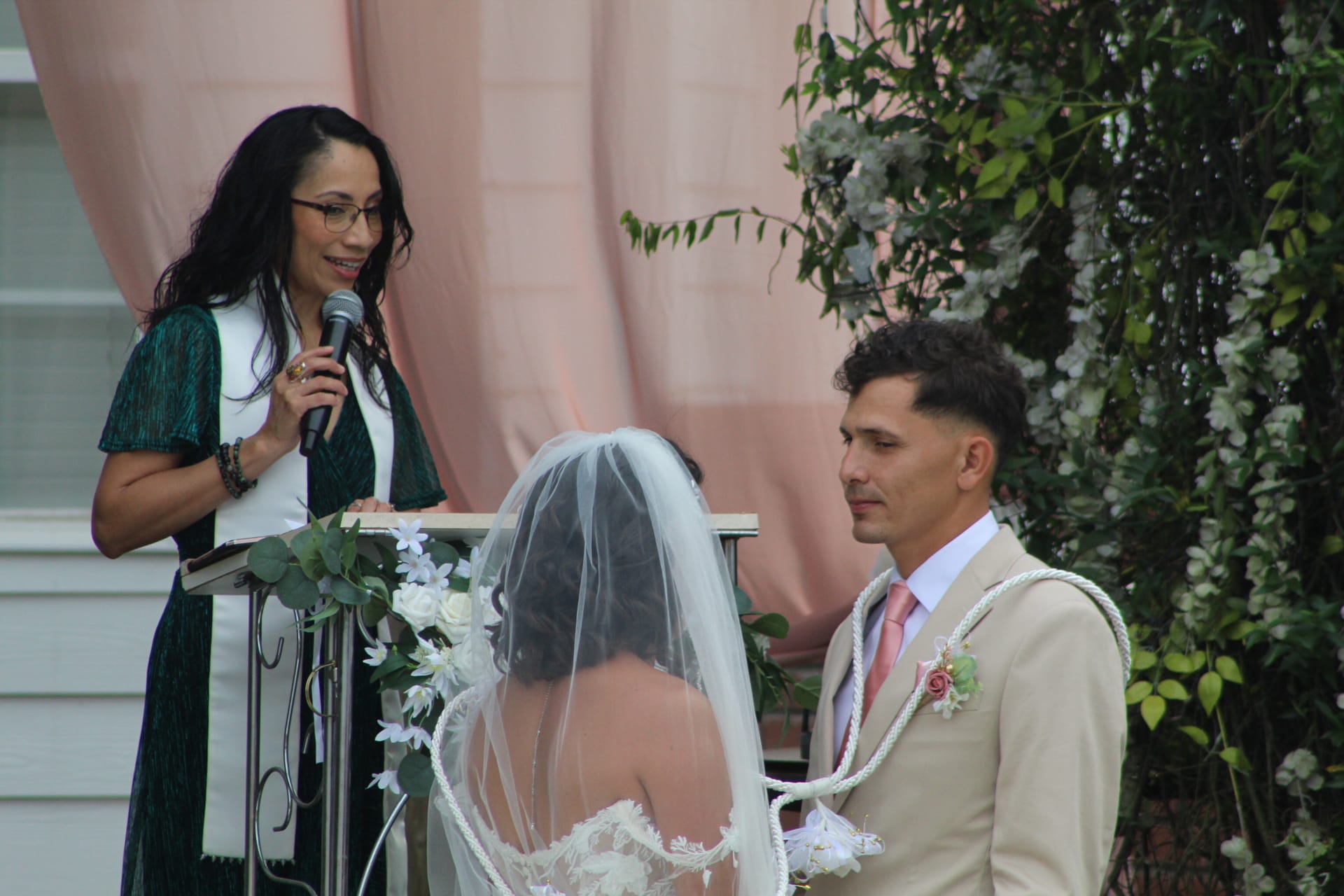 An officiant speaks at a podium while a veiled bride and suit-clad groom stand before her during an outdoor wedding ceremony at a historic estate venue decorated with white florals and soft pink drapery.