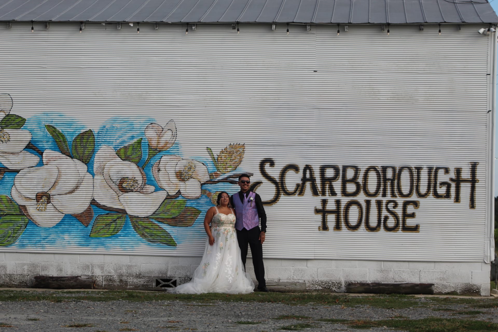 Bride in a white lace gown and groom in a black suit with purple vest pose together in front of the Scarborough House magnolia mural on the exterior barn wall.