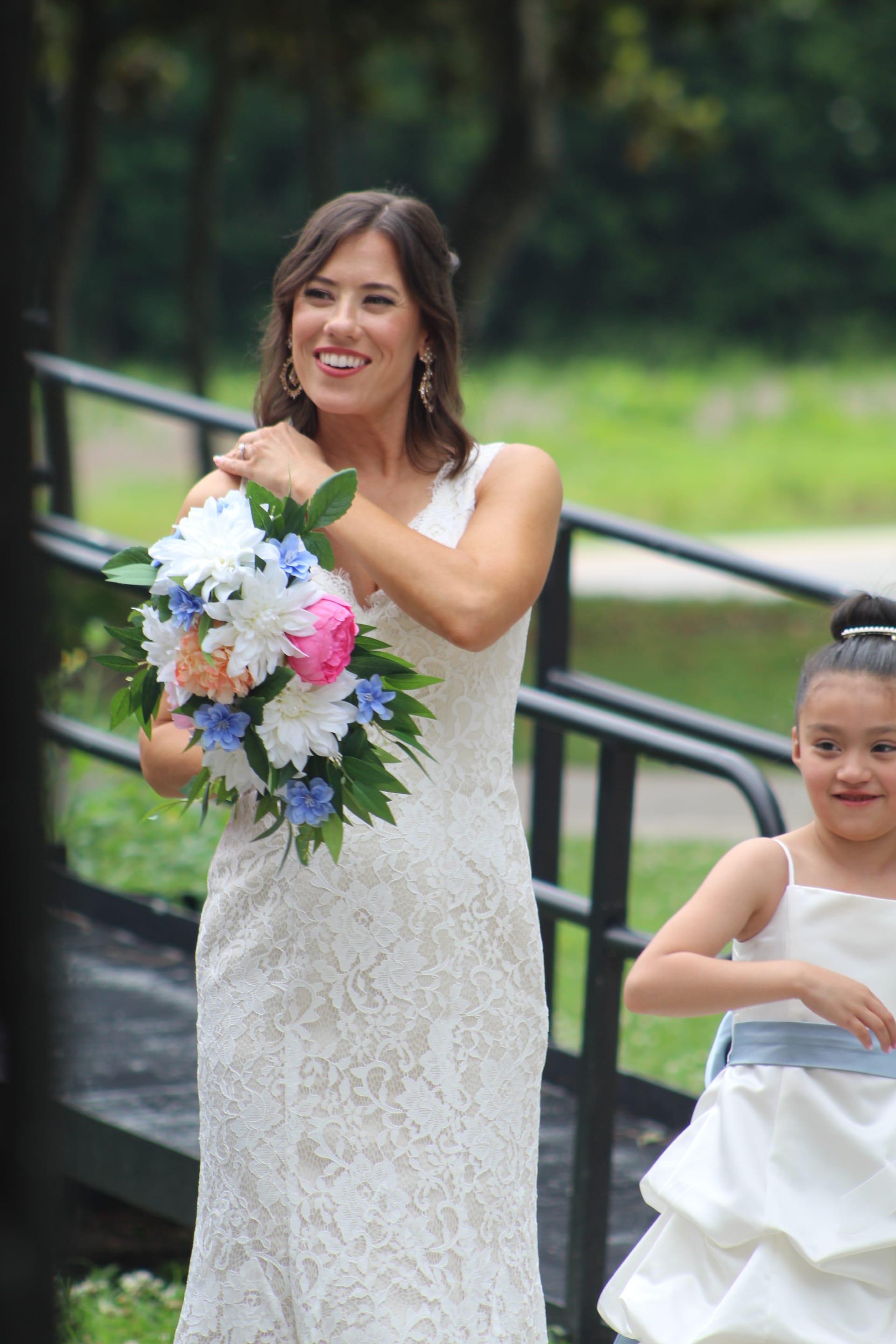 A joyful bride in a lace gown holds a colorful bouquet of pink peonies and blue wildflowers while walking alongside a flower girl in a white dress with a blue sash at an outdoor wedding venue.