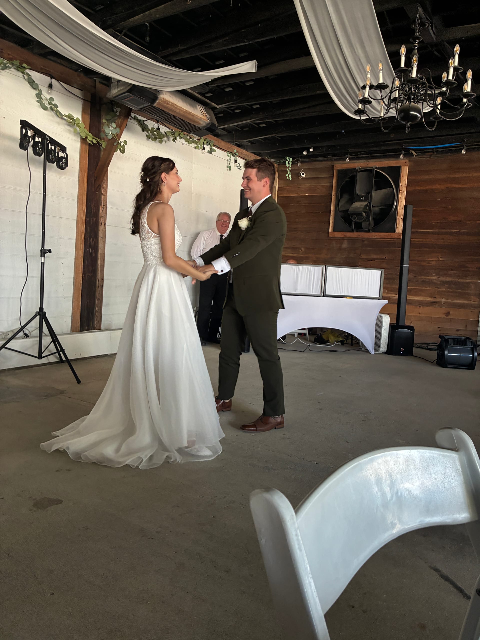 Bride in a flowing white gown and groom in an olive suit share their first dance in a rustic indoor venue space decorated with draped fabric and greenery.