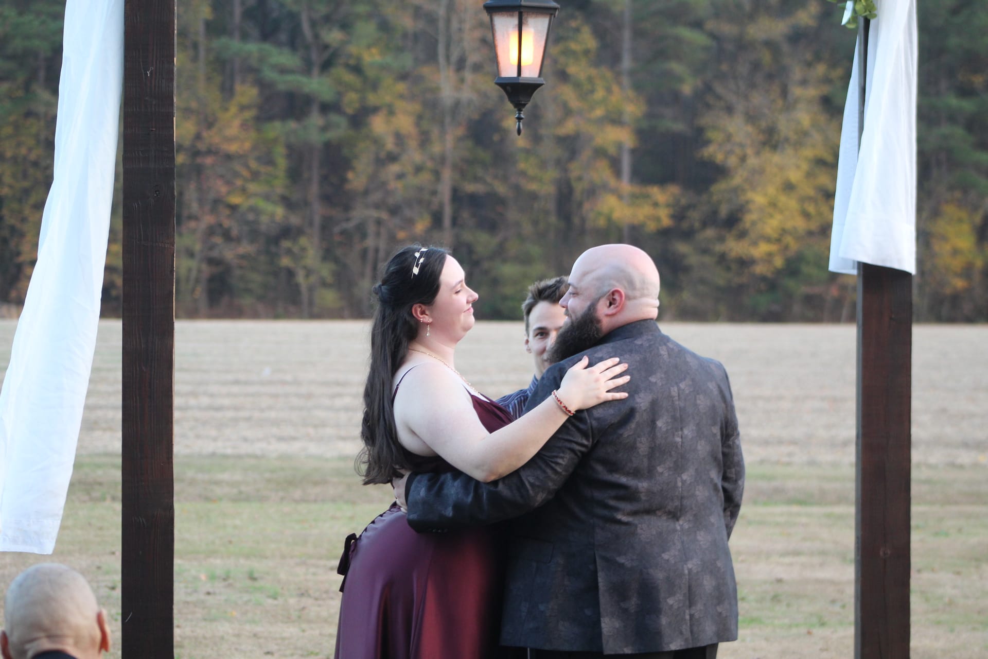 Bride in burgundy gown smiling as she embraces her bearded groom beneath a wooden arch with a glowing lantern during an outdoor autumn wedding ceremony at Scarborough House.