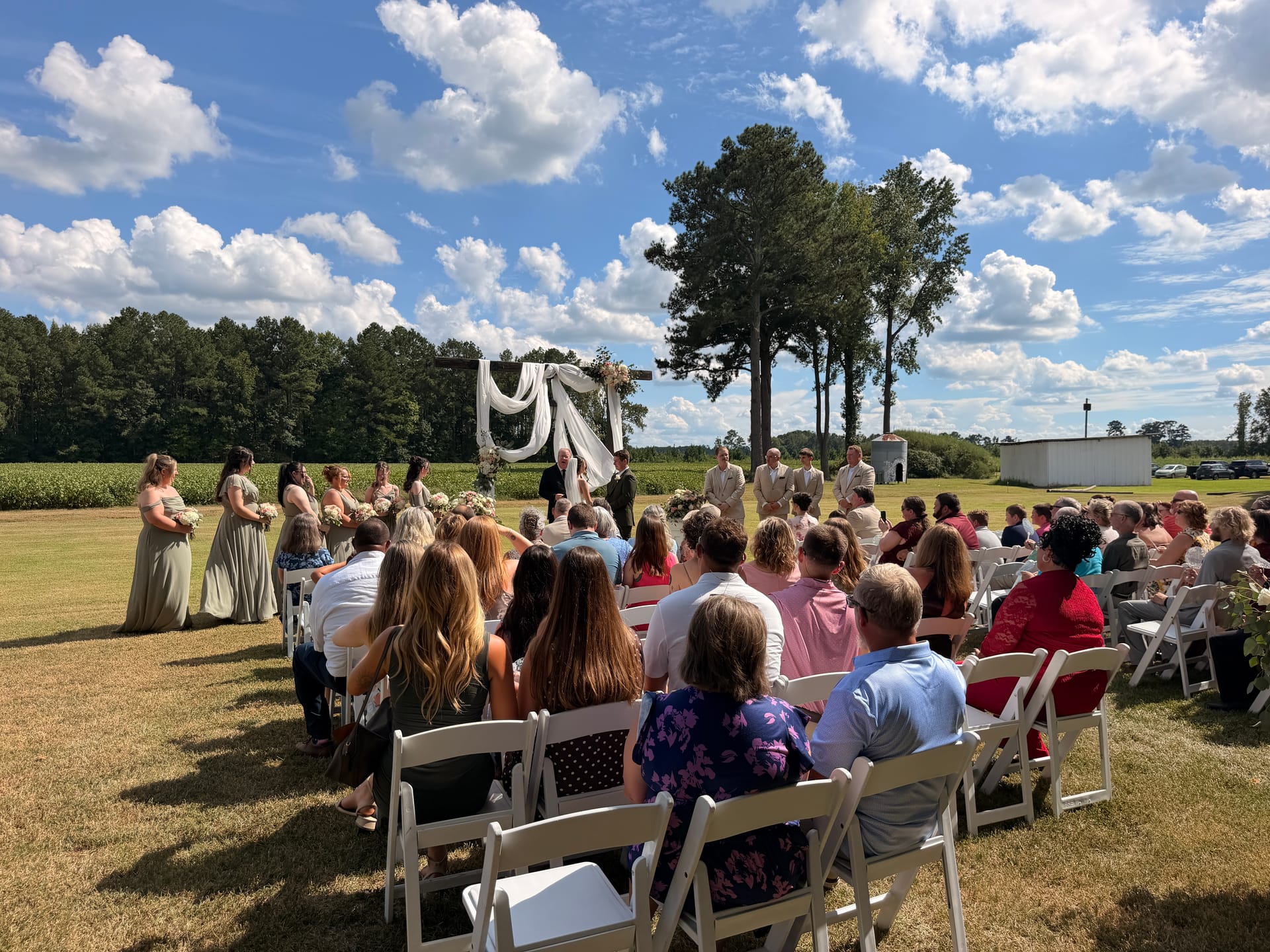 Guests seated in white garden chairs watch a wedding ceremony beneath a draped floral arch on the open ceremony lawn at Scarborough House, surrounded by farmland fields and tall trees under a blue sky.