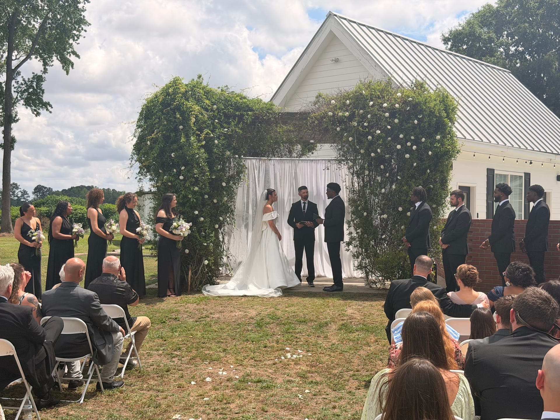 Bride and groom exchange vows beneath a lush ivy and white floral archway on the ceremony lawn at Scarborough House, surrounded by bridesmaids in black gowns and groomsmen in dark suits before seated guests.