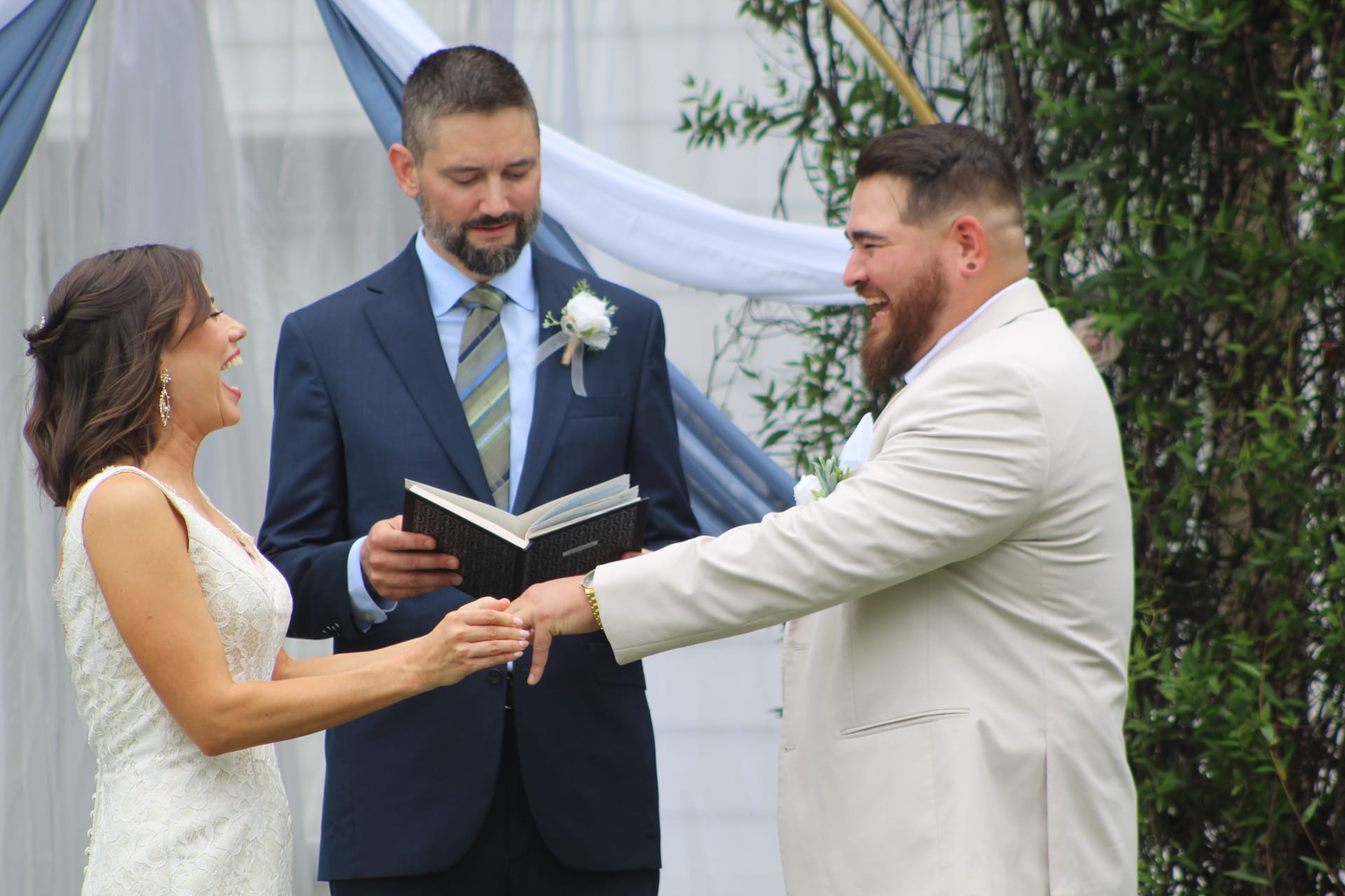 A laughing bride in a lace gown and a bearded groom in a cream suit share a joyful moment during their ring exchange while an officiant reads from a book beneath a draped blue arch at Scarborough House.