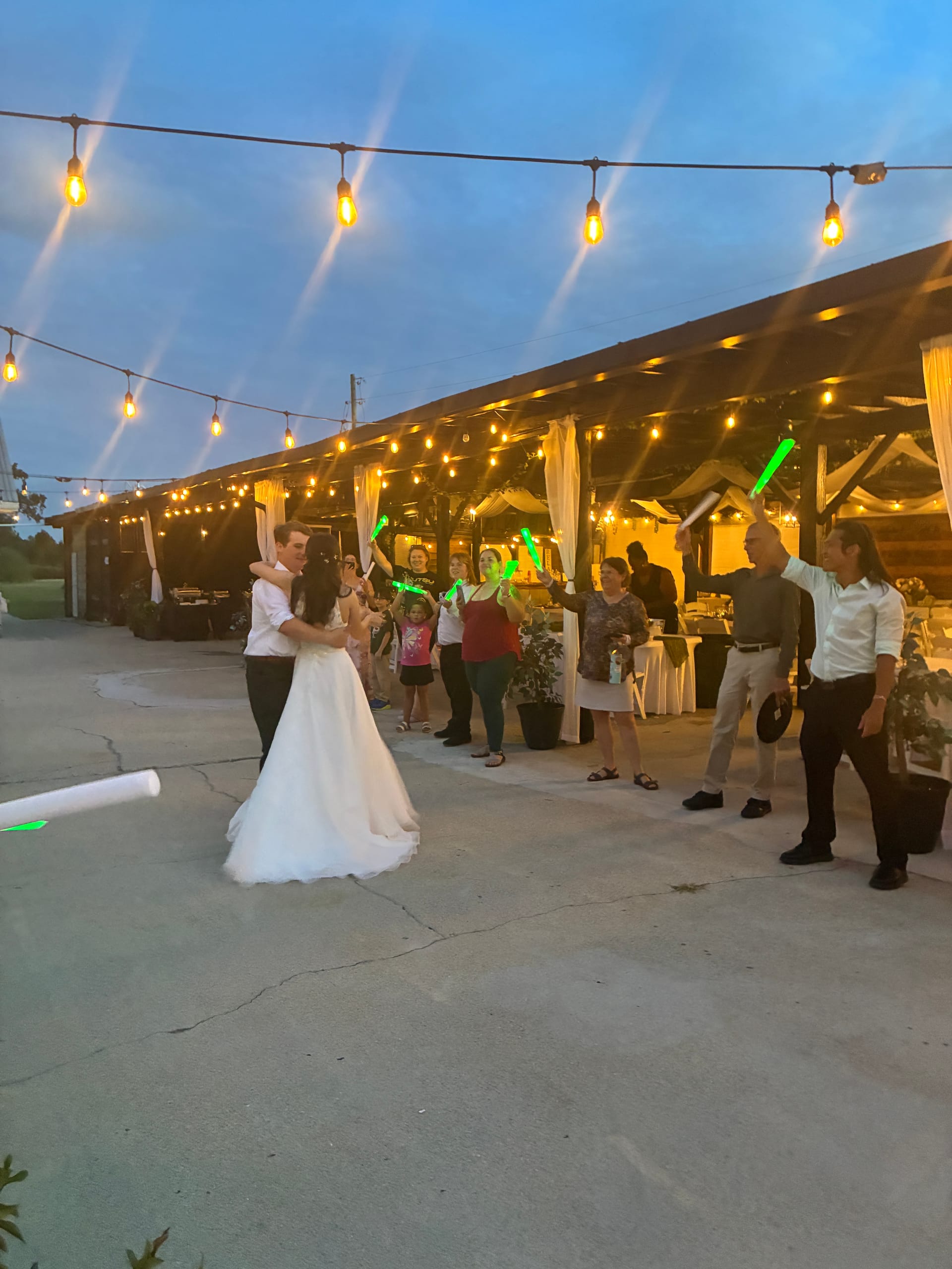 Bride and groom share their first dance on an outdoor patio at dusk as guests hold glowing green light sticks beneath warm string lights at Scarborough House wedding venue.
