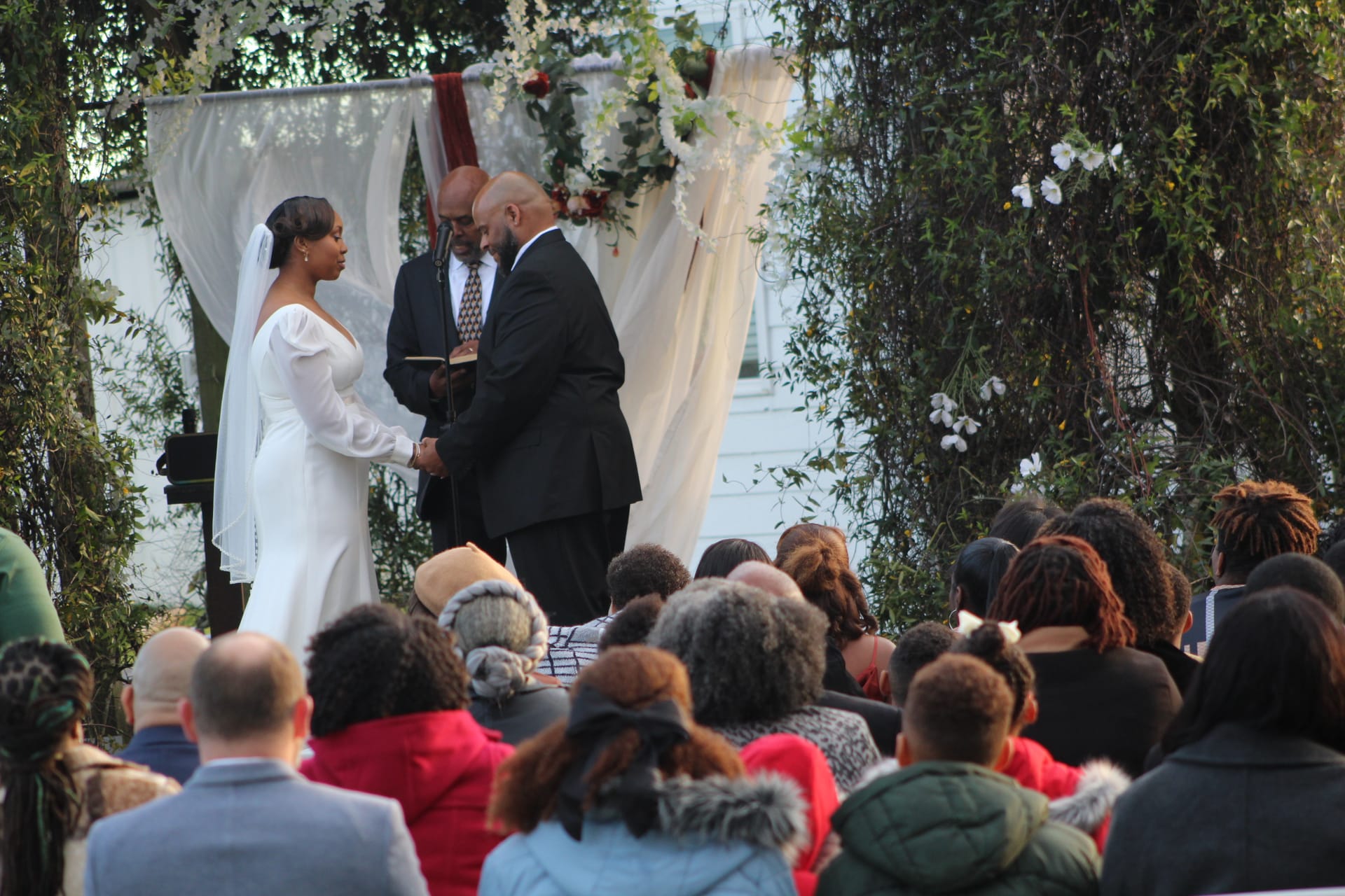 Bride and groom hold hands while exchanging vows beneath a flower-adorned arch surrounded by guests at an outdoor wedding ceremony at Scarborough House.