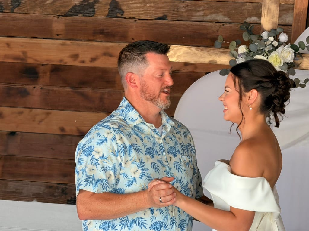 Bride in an off-shoulder white gown smiles at her groom in a blue floral shirt as they hold hands beneath a white arch adorned with white roses and eucalyptus at Scarborough House.