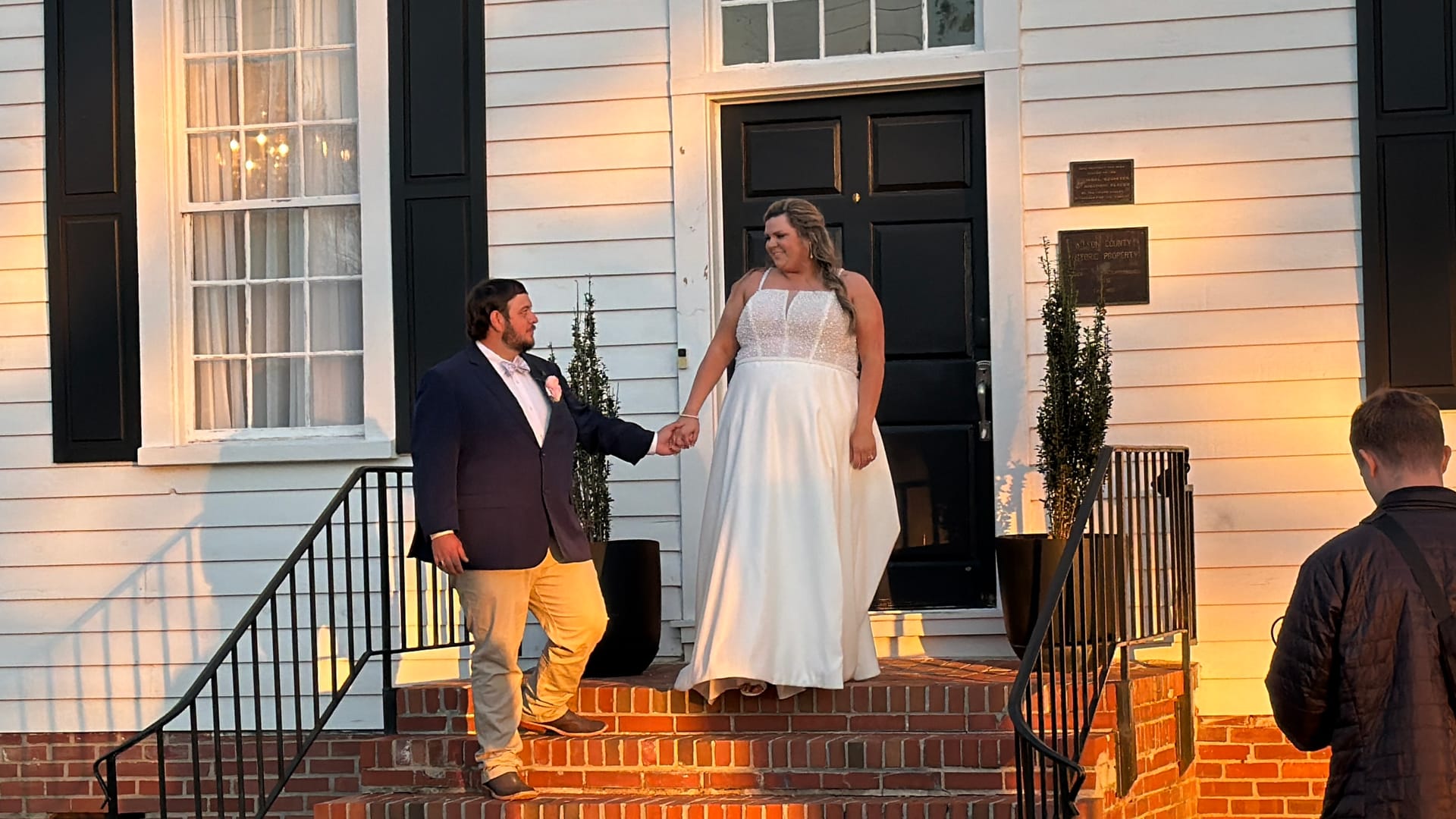 Bride in a white gown and groom in a navy blazer hold hands on the brick front steps of the 1821 Scarborough House manor at golden hour.