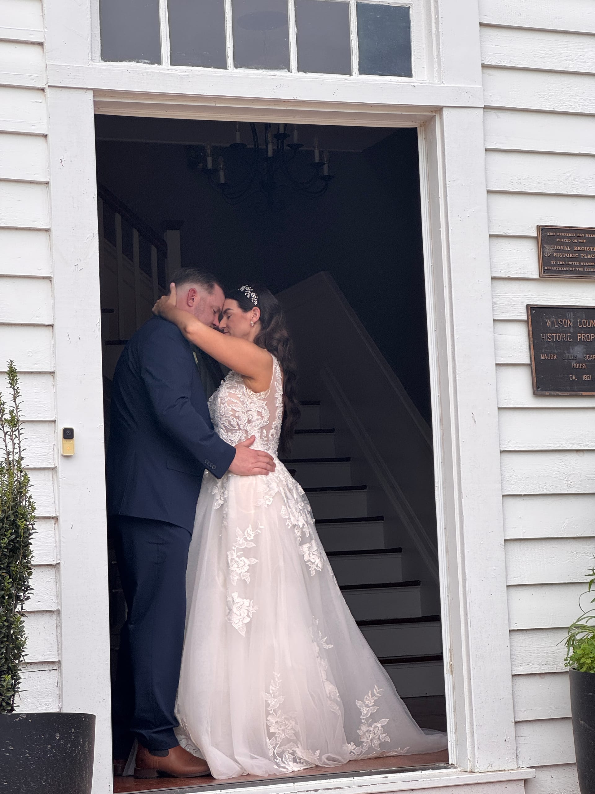 Bride and groom share a tender forehead-to-forehead embrace in the grand white doorway of the historic 1821 Scarborough House manor, the bride wearing an elegant lace ballgown with a flowing veil.