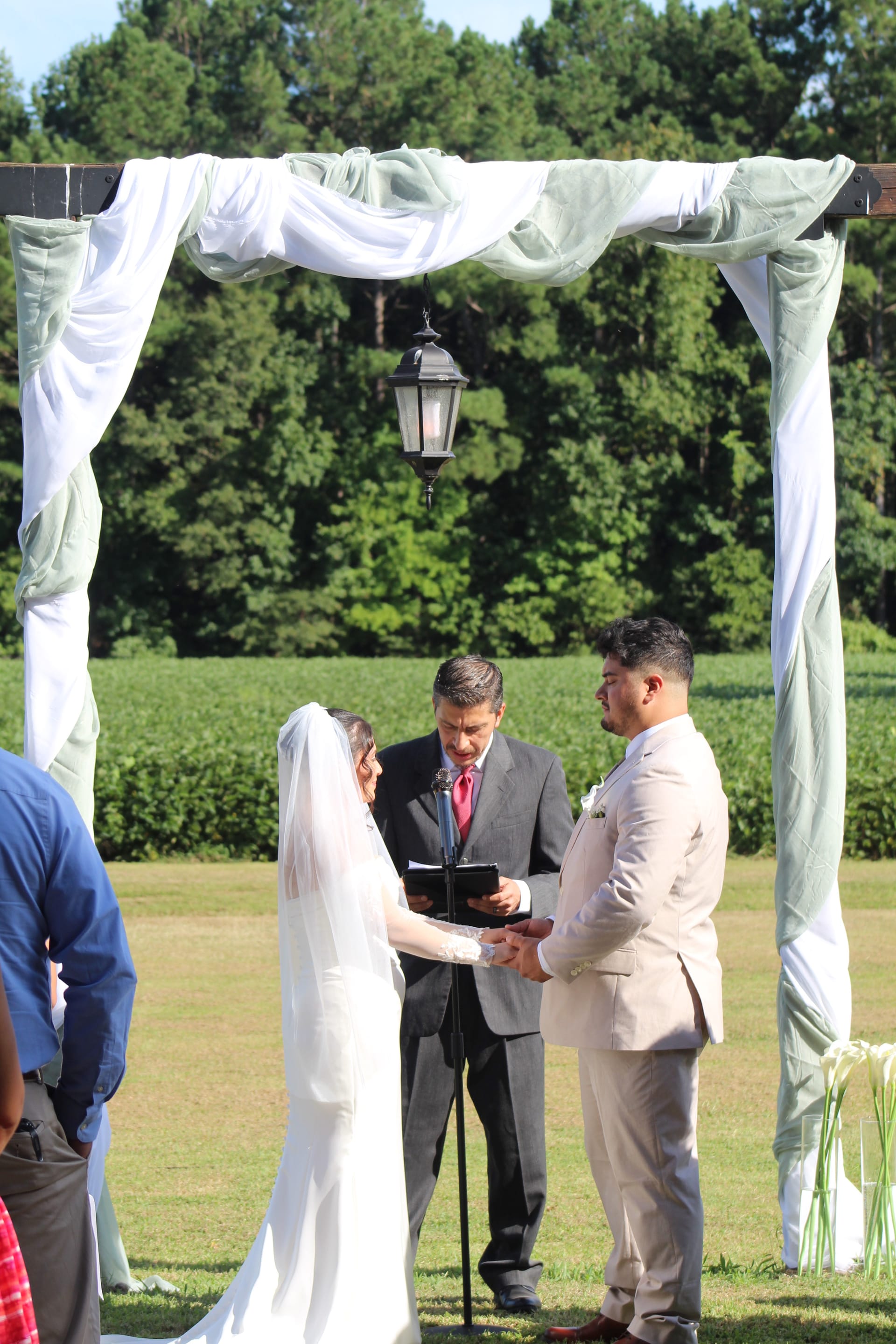 Bride and groom hold hands during outdoor wedding ceremony beneath a draped wooden arch with hanging lantern, officiant reading from a book with green farmland fields and trees in the background at Scarborough House.