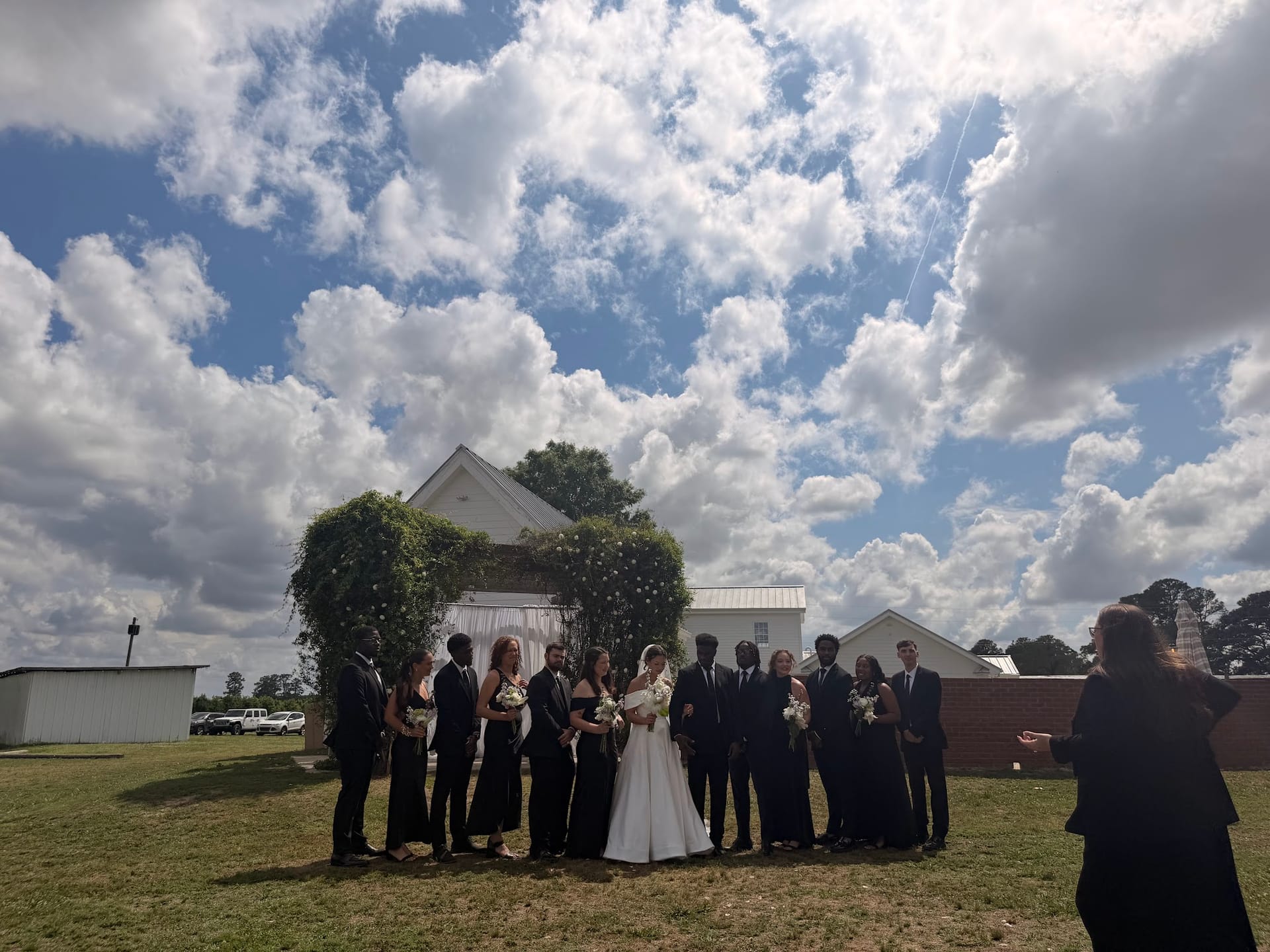 Wedding party in black attire flanks the bride and groom beneath an ivy-covered arch on the ceremony lawn at Scarborough House under a dramatic cloudy sky.