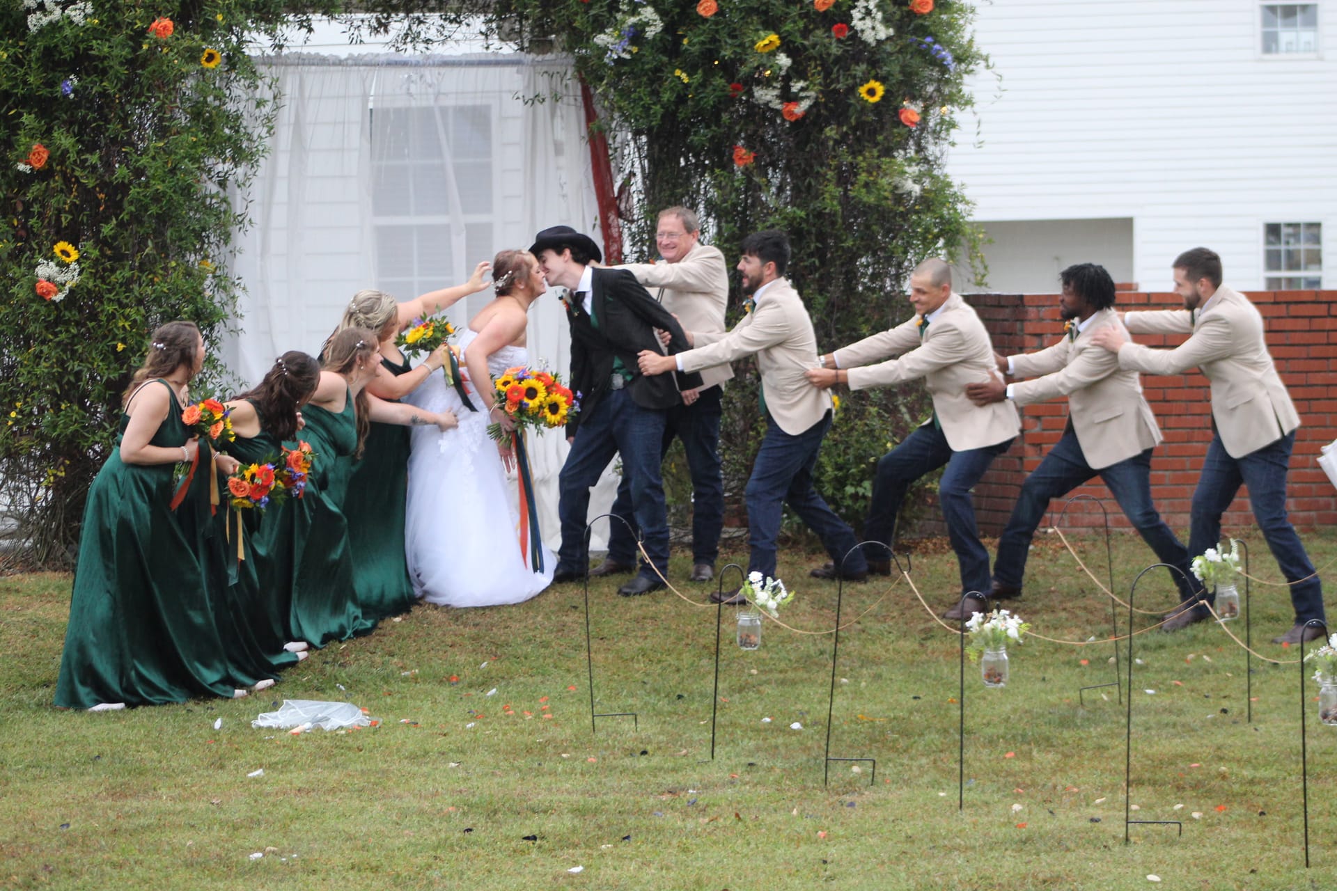 Bride and groom share a kiss while bridesmaids in emerald gowns and groomsmen in tan blazers playfully pull the couple apart in a tug-of-war pose on the ceremony lawn at Scarborough House, surrounded by colorful floral arrangements.