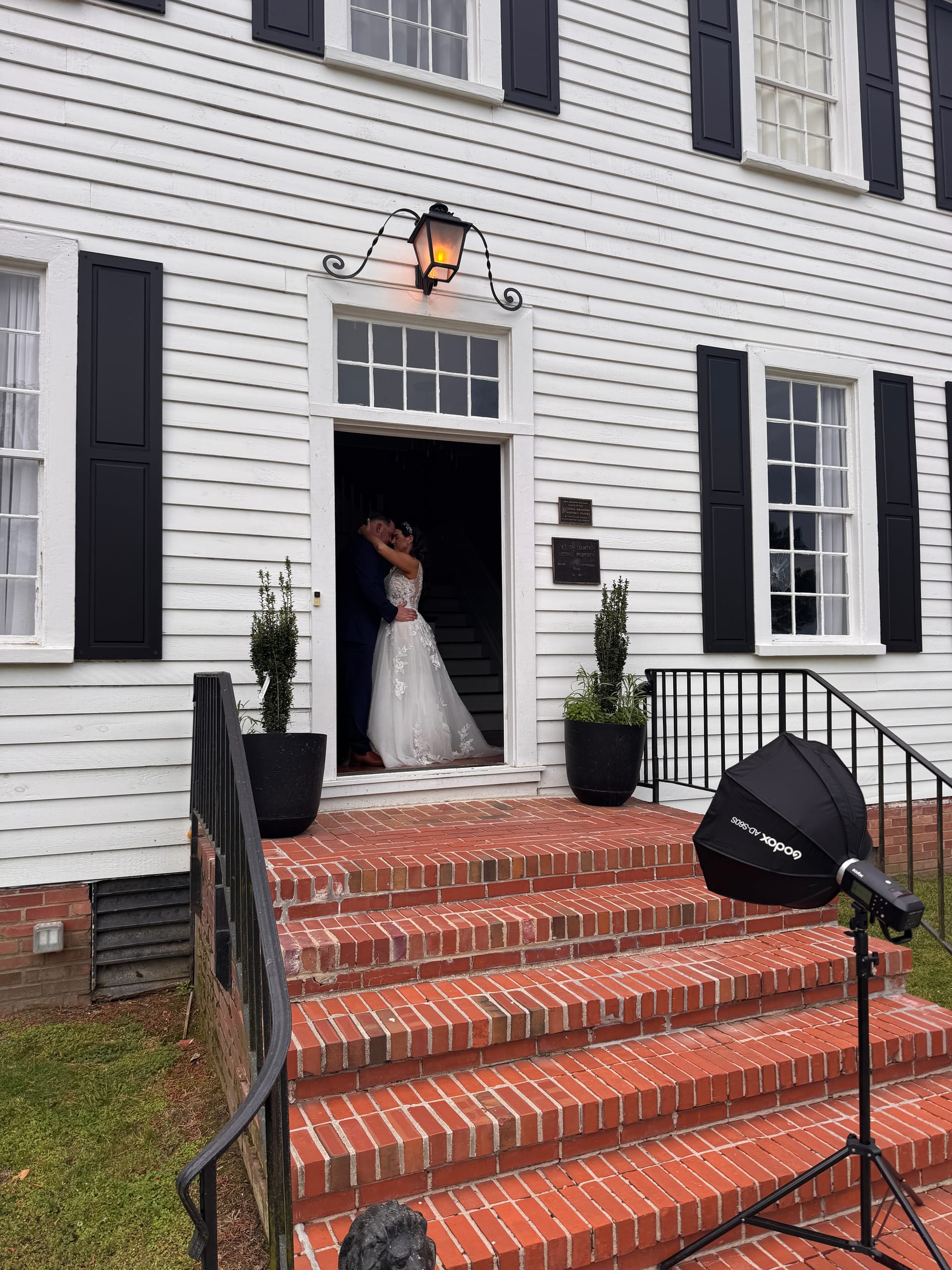 Bride and groom sharing a romantic embrace in the illuminated doorway of the 1821 Scarborough House manor, flanked by brick steps and black-shuttered windows.