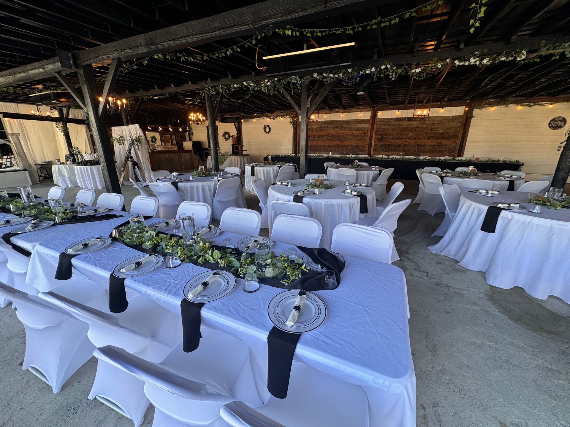Elegantly set reception tables with white linens, black napkins, silver charger plates, and lush greenery runners arranged beneath a rustic wooden pavilion with string lights and chandelier lighting at Scarborough House.