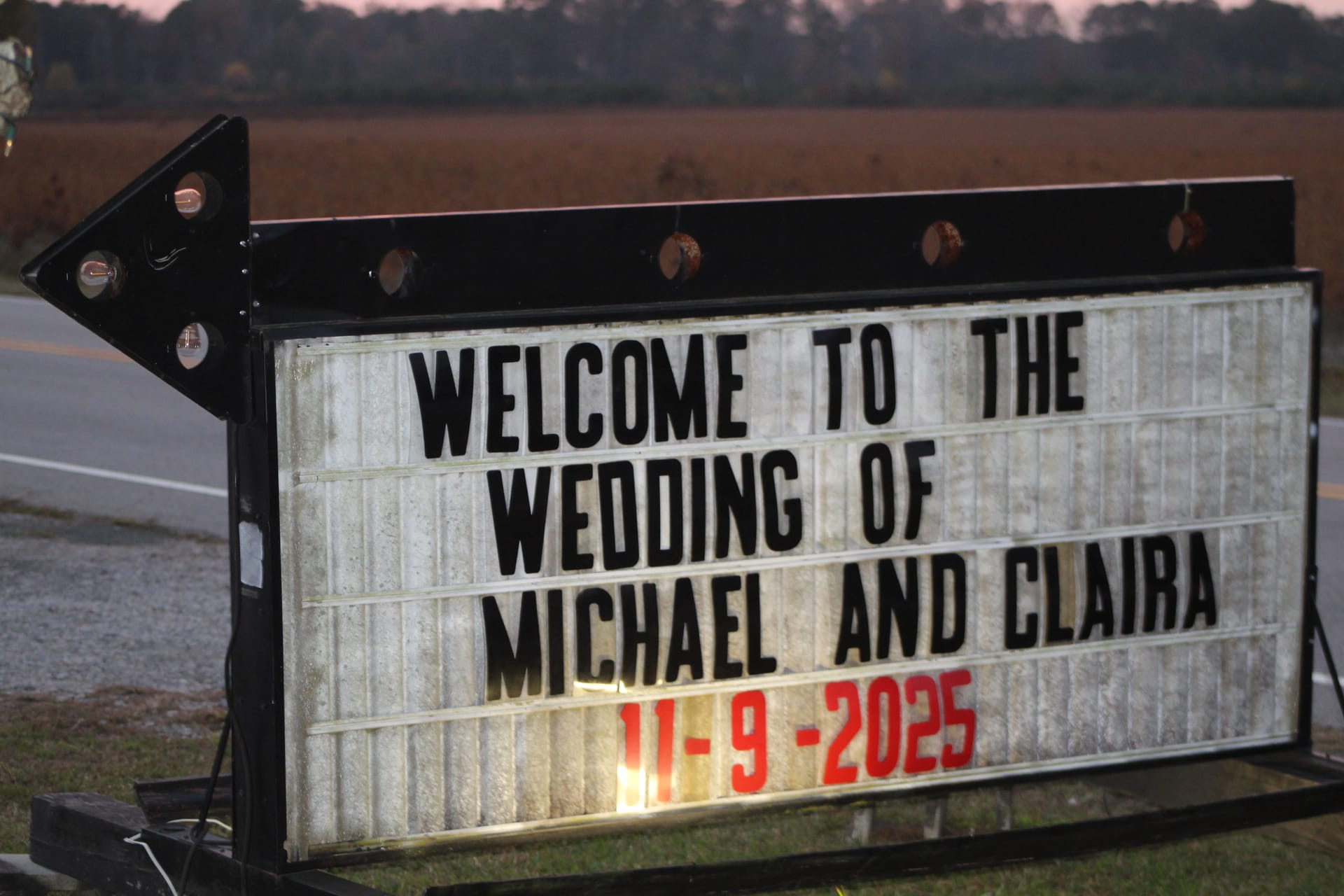 Illuminated roadside marquee sign welcoming guests to the wedding of Michael and Claira on November 9 2025 at dusk with farmland fields in the background.