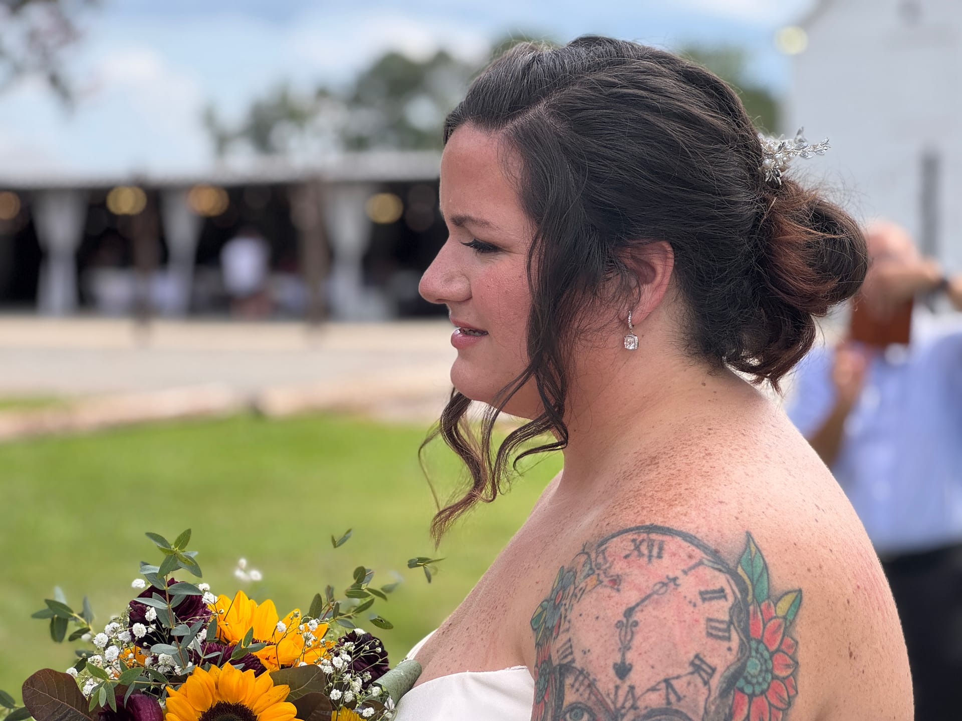 Bride in profile wearing an updo with floral hair accessory holds a sunflower and burgundy bouquet at an outdoor wedding venue, her back tattoo of a clock and flowers visible on her shoulder.