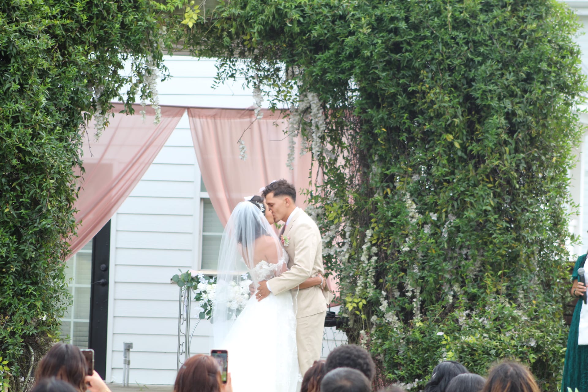 Bride and groom share their first kiss as newlyweds beneath a pink draped arch surrounded by lush greenery at Scarborough House outdoor ceremony lawn.
