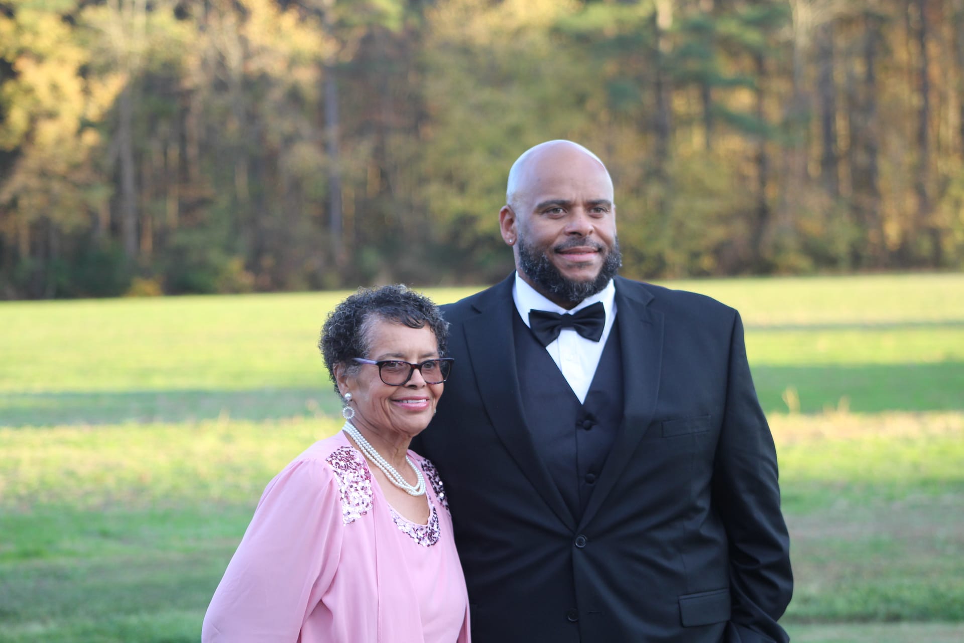 An elegantly dressed woman in pink and a man in a black tuxedo smile together on the open farmland fields of Scarborough House estate.