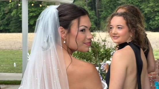 Bride in a white gown and pearl-trimmed veil glances back with a warm smile while a guest in a black dress looks on at an outdoor garden wedding venue.