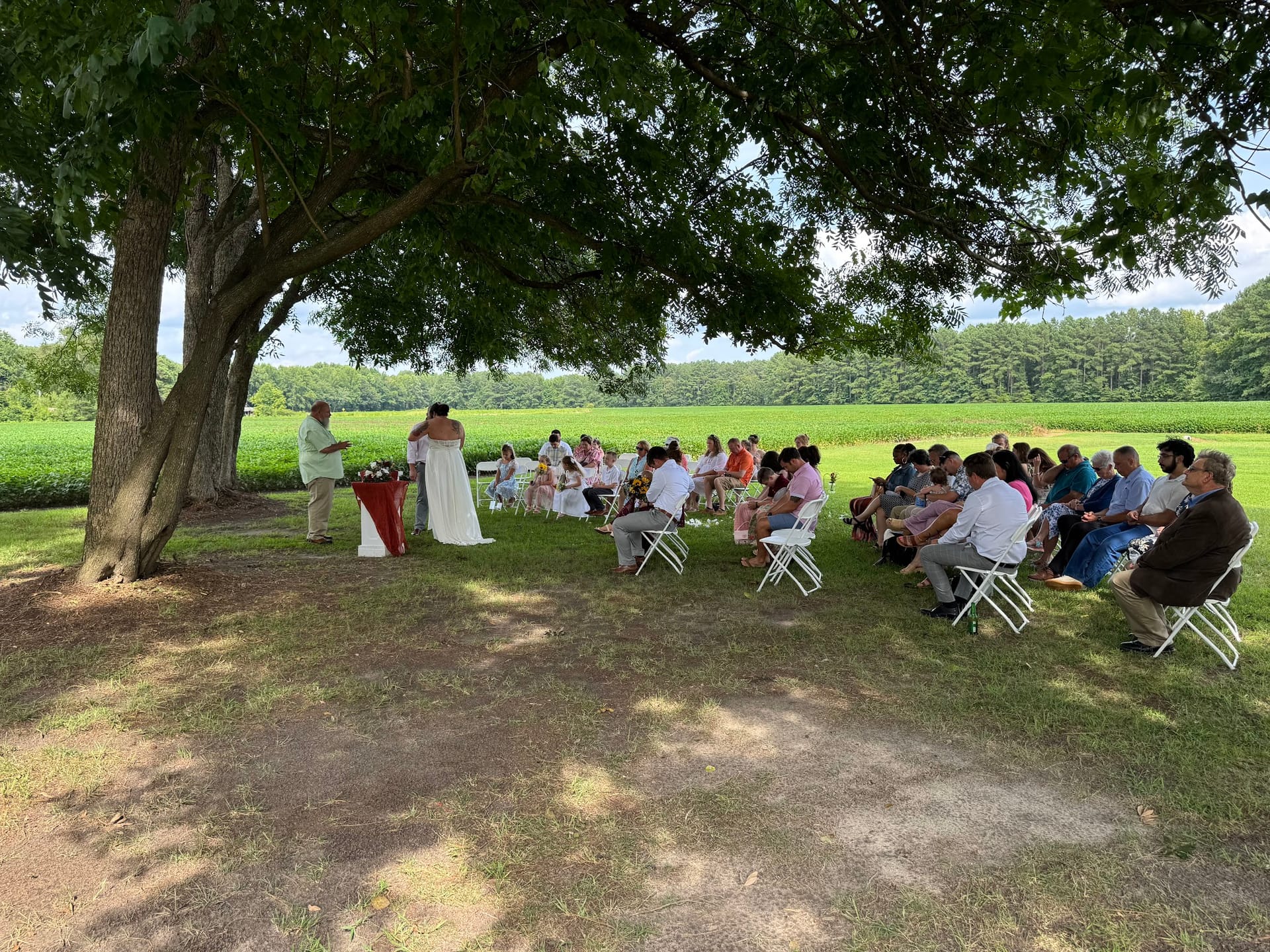 An intimate outdoor wedding ceremony held beneath a large oak tree on the ceremony lawn at Scarborough House, with guests seated in white chairs facing the couple and lush green farmland fields stretching into the background.
