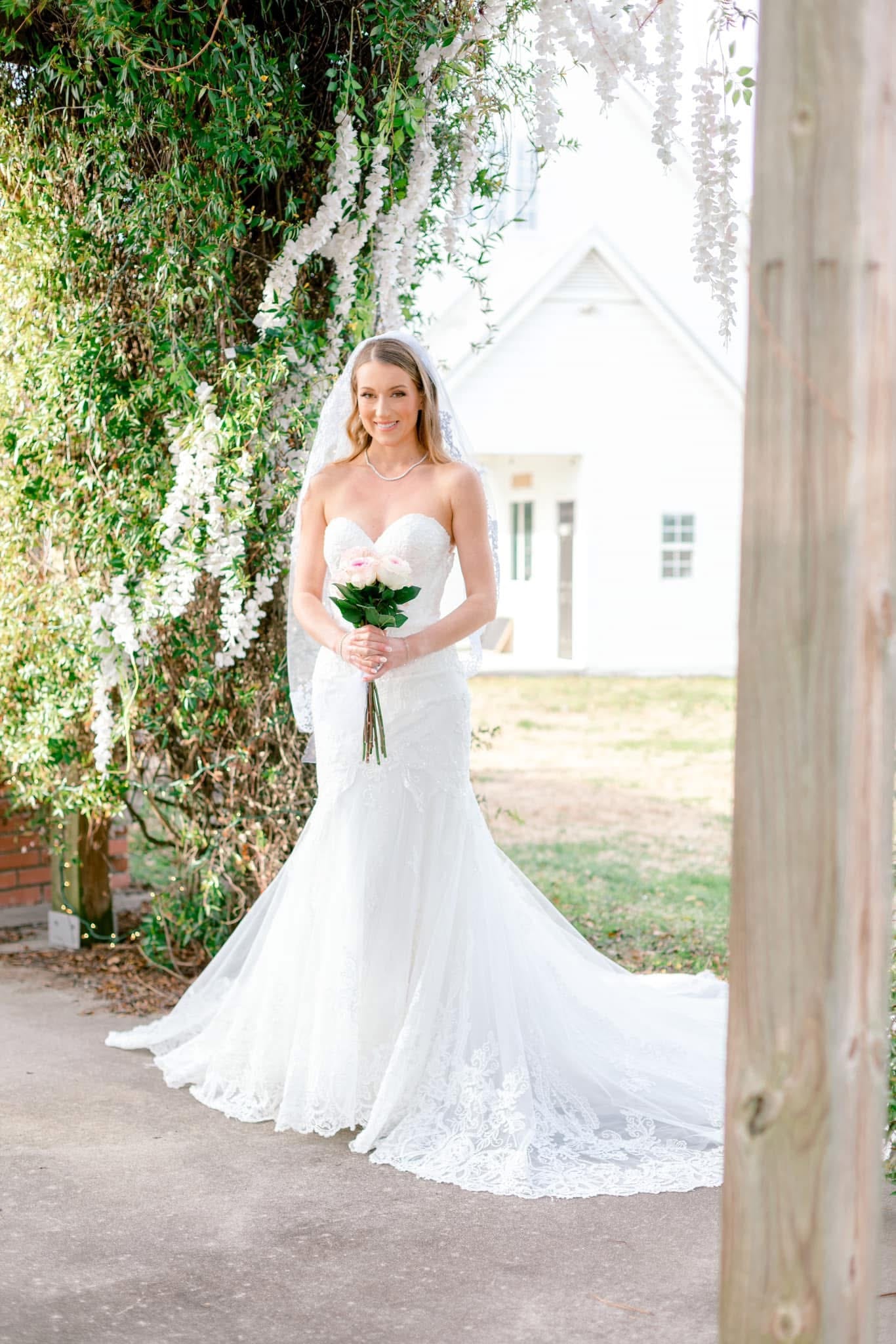 Bride in a strapless lace mermaid gown and veil holding a pink rose bouquet smiling beneath cascading white wisteria at Scarborough House historic estate.