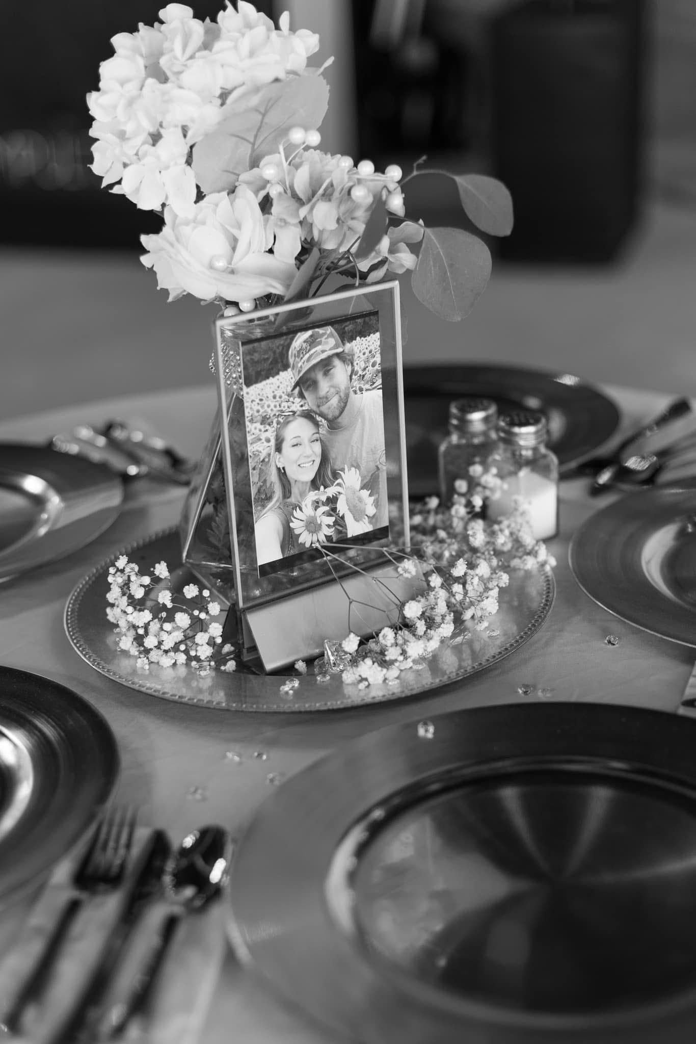 Black and white close-up of a wedding reception table centerpiece featuring a framed photo of the couple surrounded by baby's breath, white florals, and silver charger plates at Scarborough House.